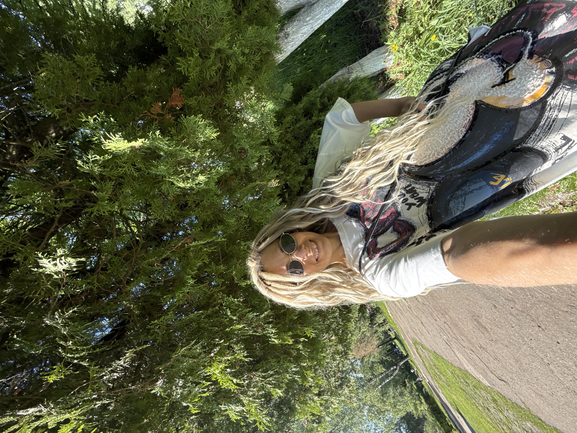 A young woman is photographed in a medium shot in what appears to be a park setting. The location is Шебунино, Russia. She is smiling, and is wearing sunglasses. Her long, blonde hair falls over her shoulder. She is wearing a white t-shirt with a graphic print. In the background, there is lush green foliage of a large tree and a street. The angle of the photo suggests it might be a selfie. The scene is well-lit, indicating a sunny day.