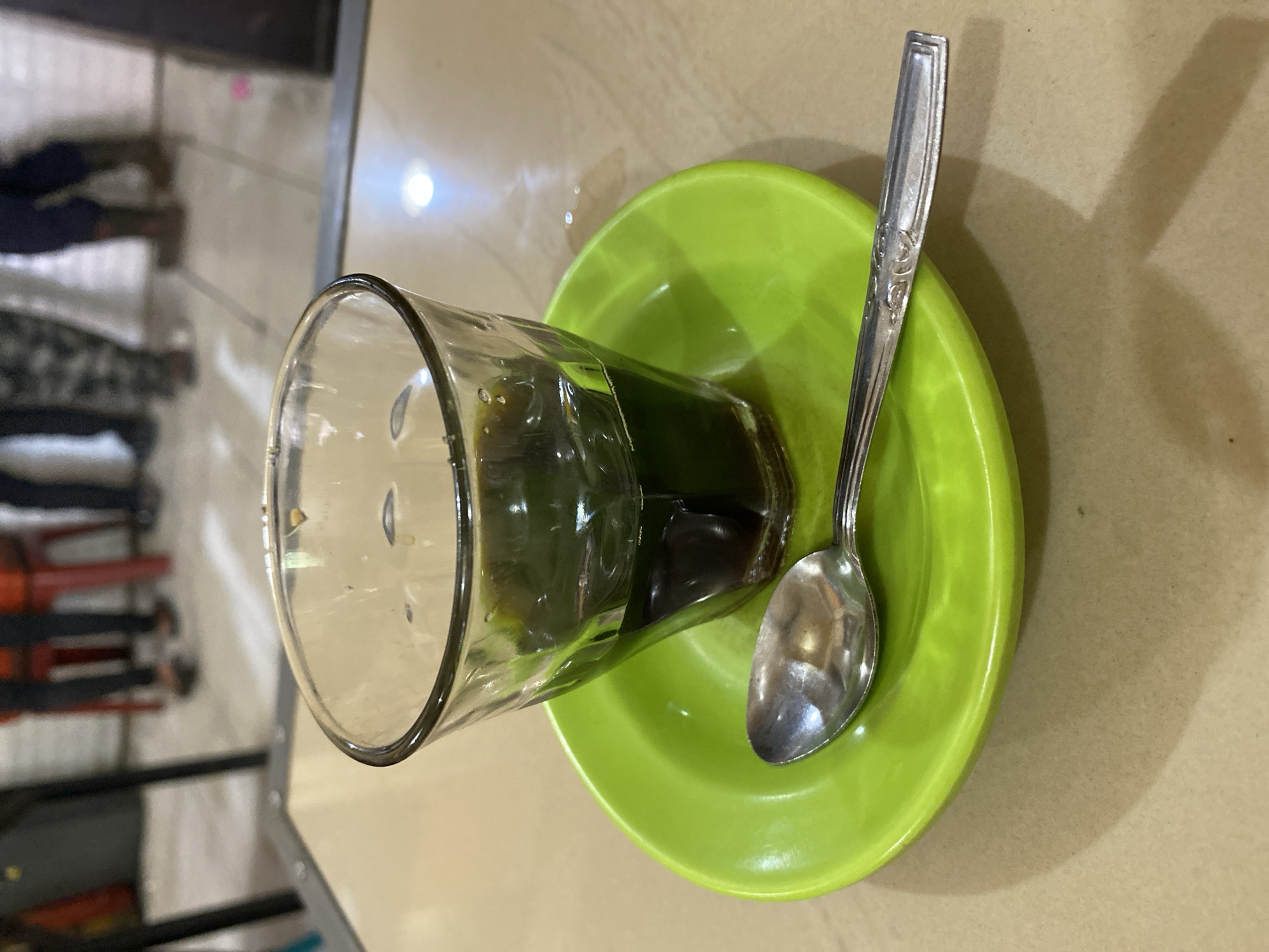 In Lhokseumawe, Indonesia, a glass of dark liquid, possibly coffee or tea, sits on a bright green saucer. A silver spoon rests on the saucer to the left of the glass. The table surface is light brown and reflects light. In the background, out of focus, are the legs of several individuals standing, some wearing dark pants and boots. They are positioned behind red chairs. The overall scene appears to be an indoor setting, possibly a cafe or restaurant.