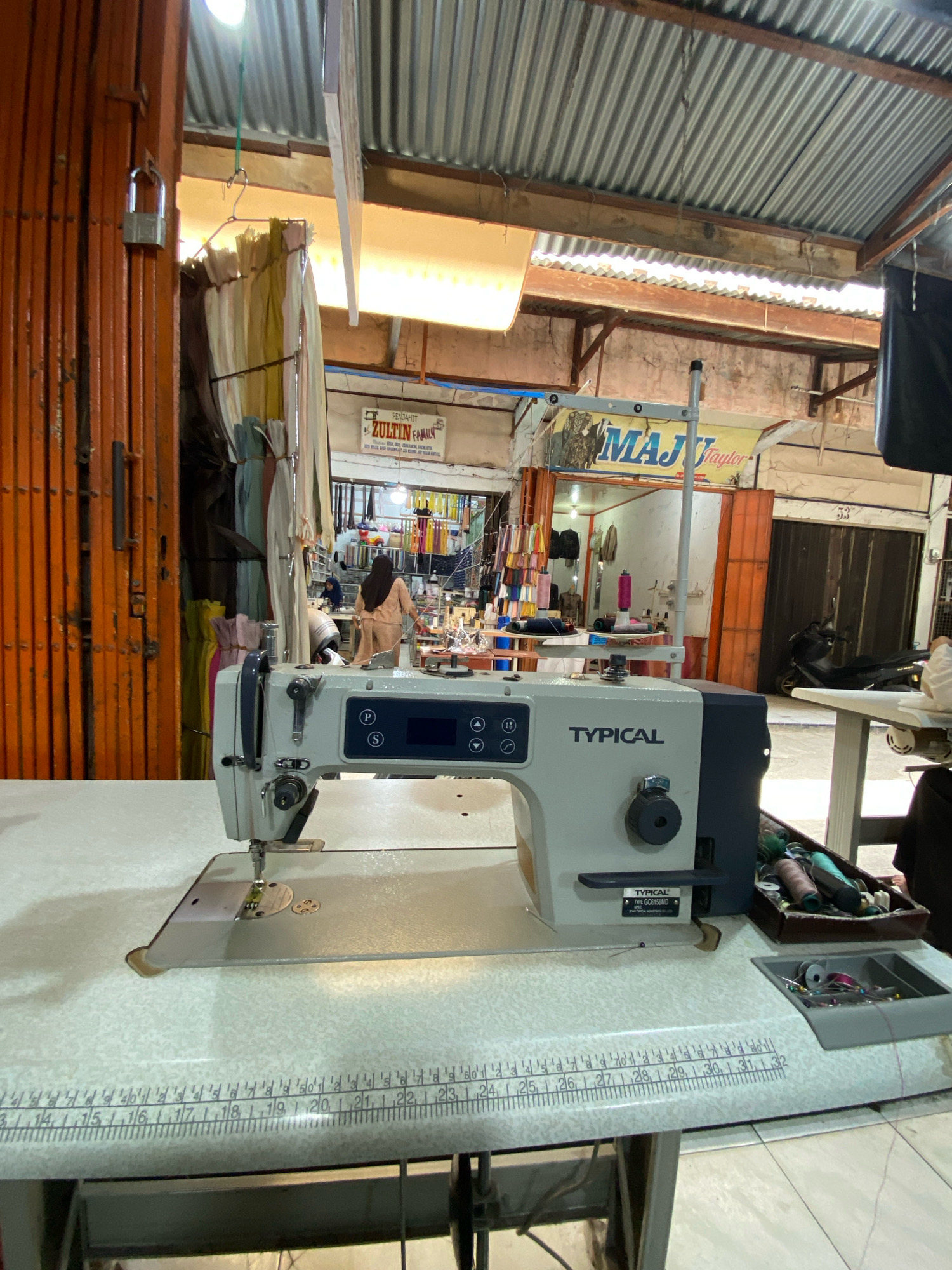 The image shows an indoor market stall in Lhokseumawe, Indonesia, featuring a sewing machine in the foreground. The stall appears to be a tailor's shop, indicated by the presence of fabric rolls, spools of thread, and a sewing machine. A woman wearing a hijab and a beige outfit is visible in the background, likely working at the stall. The market setting is characterized by corrugated metal roofing and orange metal shutters, suggesting a functional rather than decorative setup. The sewing machine is a "TYPICAL" brand, Model GC6150MD, and is positioned on a patterned table with a built-in measuring tape. A wooden box filled with colorful spools of thread and a tray with needles and thread are also visible on the table. The overall scene depicts a working environment within a commercial district of the city.