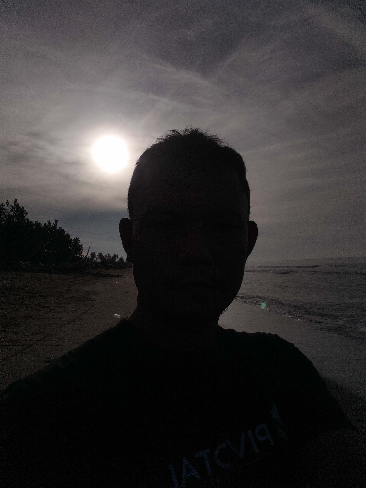 The image captures a silhouetted male figure on a beach in Curehtunong, Indonesia. The individual, facing the camera, is positioned centrally in the frame. The sun, a bright, overexposed white circle, is situated in the upper-left quadrant, directly behind the subject's head, acting as a strong backlight. The sky is predominantly overcast with grey clouds, exhibiting some textural variations. To the subject's left, a sandy beach extends towards a dense line of trees, including what appear to be palm trees, with vague forms resembling small boats visible near the foliage. To the subject's right, the ocean is visible, with minor waves breaking on the shoreline. The person's face, hair, and upper torso are rendered in complete silhouette, obscuring any distinct features. A reversed reflection of text, possibly a logo, is faintly discernible on the front of the subject's dark shirt. The scene is dominated by dark tones and silhouettes due to the direct backlighting.