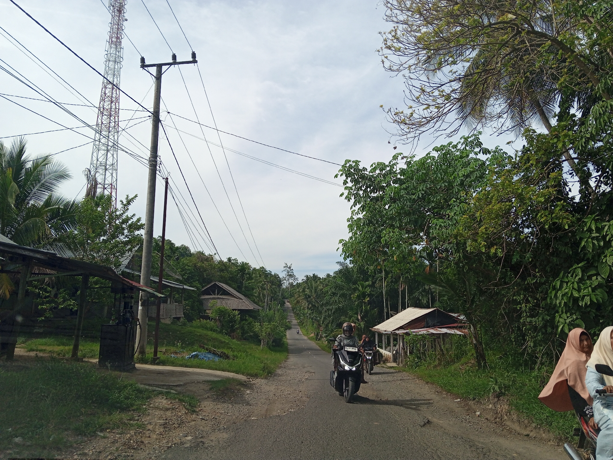 The image depicts a rural road scene in Meunasahbeunot, Indonesia, under an overcast, light grey sky. A two-lane asphalt road, exhibiting cracks and gravel patches, extends into the background, gently ascending and curving to the left.
In the immediate foreground, a person wearing a helmet and a grey long-sleeved top rides a black scooter, identifiable by the license plate "BL 3948 BKQ," directly towards the viewer. A passenger is seated behind them. Behind this scooter, another person on a dark scooter also approaches. On the far right edge of the frame, two individuals are partially visible on a third scooter; one wears a light brown hijab, and the other a cream-colored hijab. Further in the distance, a small, dark object, likely another scooter, is visible on the road.
To the left of the road, a simple, open-sided structure with a corrugated metal roof provides shelter over a dark, rectangular object, possibly a rudimentary fuel dispenser. Behind this, several modest houses with corrugated metal roofs are visible amidst dense green foliage. A tall, red and white lattice-patterned communications tower, equipped with multiple antennas, dominates the left skyline, alongside a concrete utility pole from which numerous black power lines crisscross the sky.
On the right side of the road, a house with a light-colored corrugated metal roof is nestled among abundant tropical vegetation. The entire scene is characterized by lush greenery, including numerous palm trees and other broad-leafed trees that line the road and extend into the surrounding landscape. Grass and weeds grow along the road shoulders.
