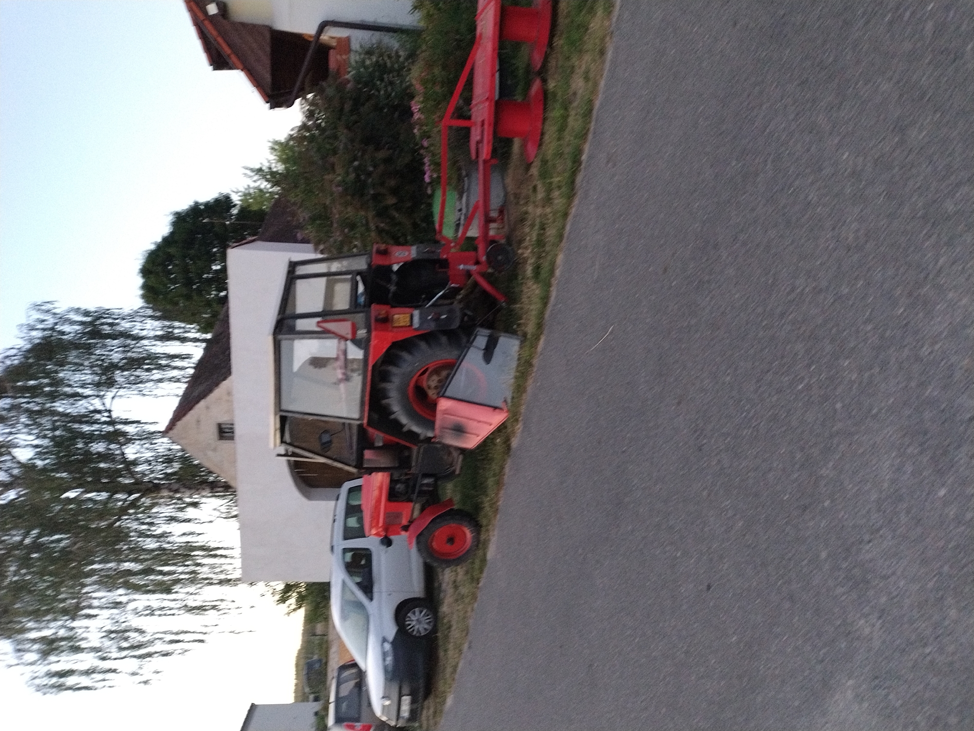 In the village of Dobev, Czechia, a rural scene unfolds with a red tractor as the central subject. The tractor, positioned on a grassy area adjacent to an asphalt road, is equipped with a trailed implement, likely a mower. To the left of the tractor, a light-colored van is parked, partially obscured by the tractor. Behind these vehicles, a white building with a dark-tiled roof stands, suggesting a residential or farm property. The background is characterized by a mature tree with drooping branches, and a clear sky. The overall impression is one of a quiet, possibly late afternoon, rural setting.