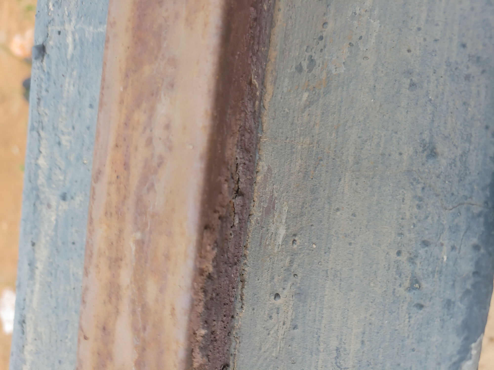 The image shows a close-up, eye-level view of a weathered, rusty metal surface adjacent to a lighter-colored, possibly concrete or painted surface. The rust appears as horizontal bands of reddish-brown and tan, with some darker, almost black areas where corrosion is more pronounced. The lighter surface below has a rough texture and is speckled with small dark spots and some lighter, rougher protrusions. The lighting suggests an outdoor environment, but no specific city or location details are visible. The focus is entirely on the texture and material of the surfaces.