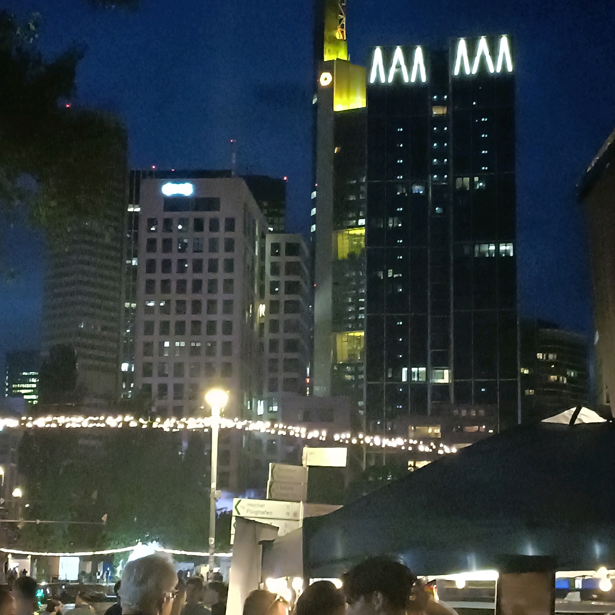 The image captures a nighttime view of the Frankfurt skyline, focusing on modern skyscrapers adorned with illuminated signage. In the foreground, a group of people are gathered, partially obscuring the lower portion of the scene. They appear to be casually interacting, with some individuals' faces visible. Above them, string lights create a festive atmosphere, illuminating trees and pathways.
Dominating the midground and background are several prominent buildings. On the far left, a tall, slender skyscraper with a grid-like facade is visible, with lights illuminating some of its windows. To its right, a lighter-colored building with a similar grid pattern stands, featuring a distinct illuminated logo. The central focus is on a cluster of darker skyscrapers, one of which is dramatically lit with a vibrant yellow section near its peak, featuring what appear to be illuminated numbers or symbols. The tallest of these dark skyscrapers is topped with large, bright white illuminated letters that spell "MAM". Additional windows on these buildings are lit, suggesting occupancy. In the far right, a portion of another building with curved architecture is visible. The sky is a deep blue, characteristic of nighttime. Signage indicating directions, including "Höchst Flughafen" (Höchst Airport), is mounted on poles near the foreground gathering, providing a specific clue to the location being a public area within Frankfurt, likely near a transportation hub or a well-known thoroughfare. The overall impression is of a dynamic urban environment at night, with a blend of architectural grandeur and social activity.
