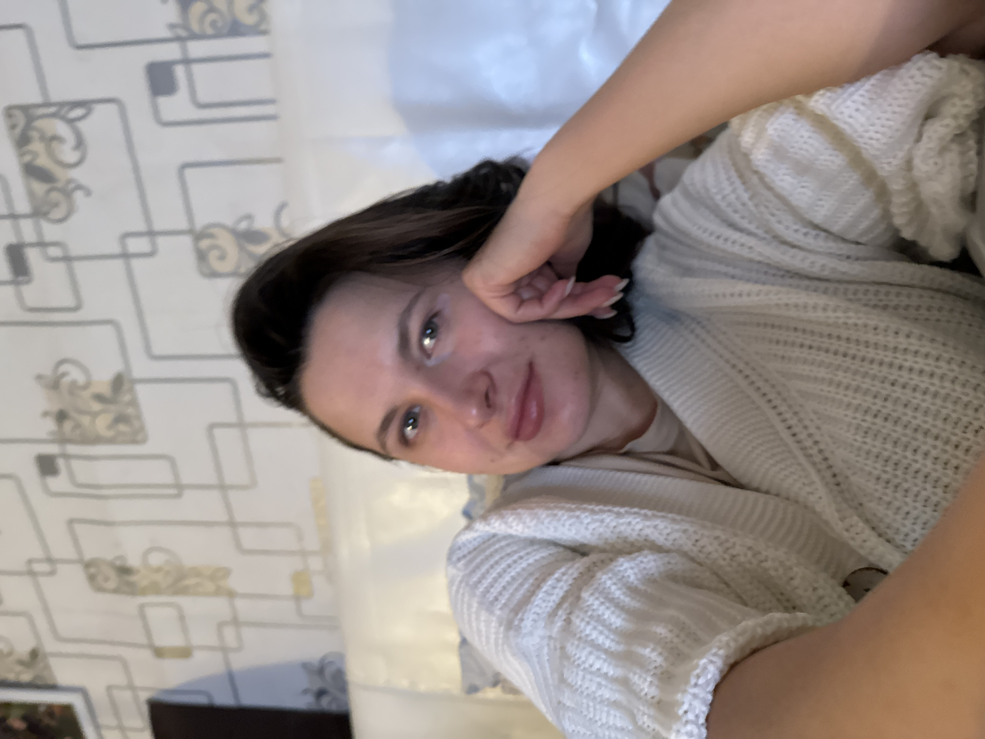 A person with dark hair and fair skin is lying on a bed. They are wearing a white, cable-knit sweater. The person's left hand is touching their face. The background includes a wall with a geometric wallpaper pattern and a white, sheet-like material. The location is Unknown, Unknown.