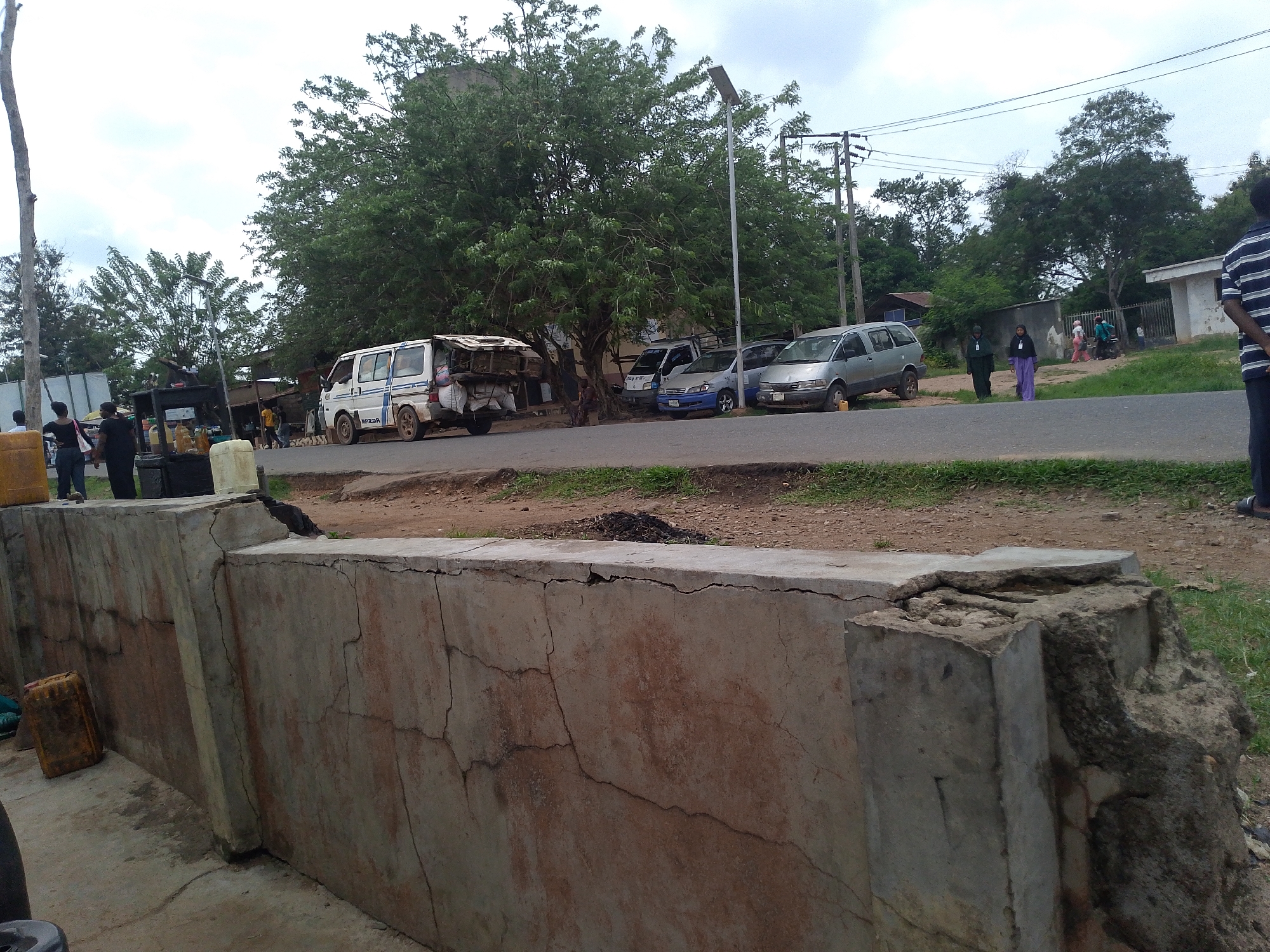 The image shows a street scene in Ipetumodu, Nigeria. A cracked, concrete wall with significant damage is in the foreground. Beyond the wall, a road runs horizontally across the frame. Several vehicles are parked along the road, including a white van with items loaded on its roof rack, a silver car, and a blue car. People are walking and standing near the vehicles and along the roadside. A large tree dominates the center of the image, providing shade to the parked vehicles. Utility poles and wires are visible in the background. The sky is overcast.
