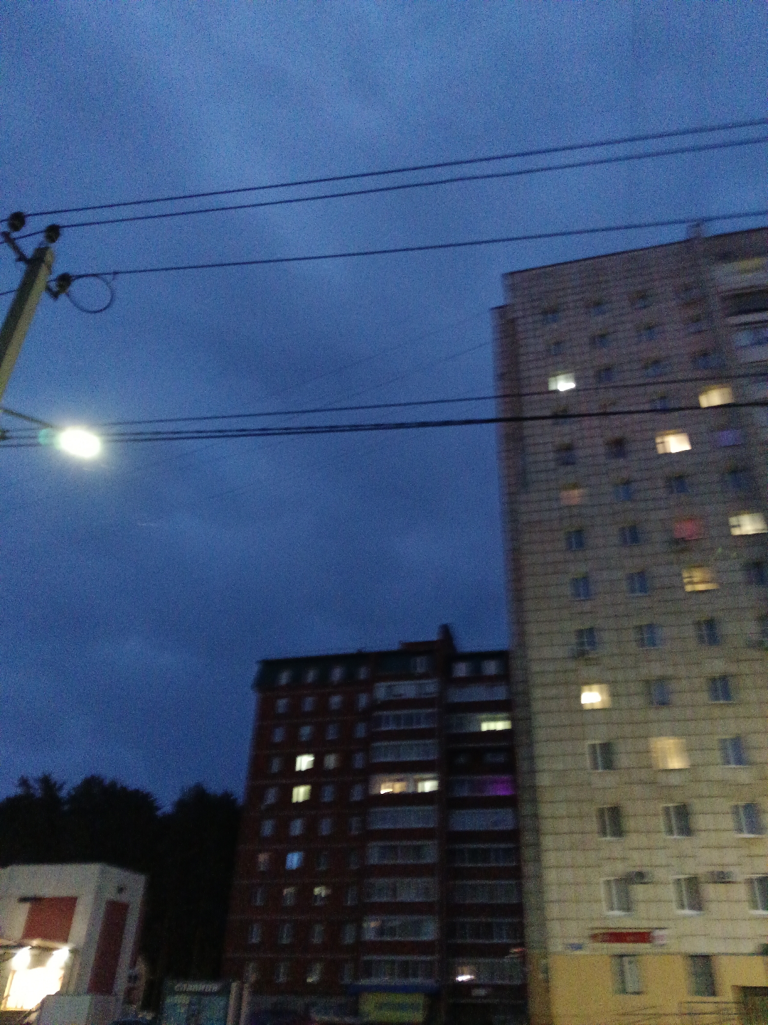 The image is a nighttime street-level photograph captured in the city of Chaikovsky, Russia. In the foreground, a power pole with a street light is visible on the left side of the frame, illuminating the dark blue sky. Three parallel power lines stretch across the upper portion of the image. Two apartment buildings dominate the mid-ground and background. The building on the right is taller and lighter in color, with numerous windows, some of which are illuminated. To the left of this building is a shorter, darker red brick apartment building, also with illuminated windows. The very bottom of the frame shows a small section of a light-colored building with red accents, likely a storefront, and the tops of dark trees can be seen between the buildings and the sky. The overall scene depicts an urban environment at dusk or night.
