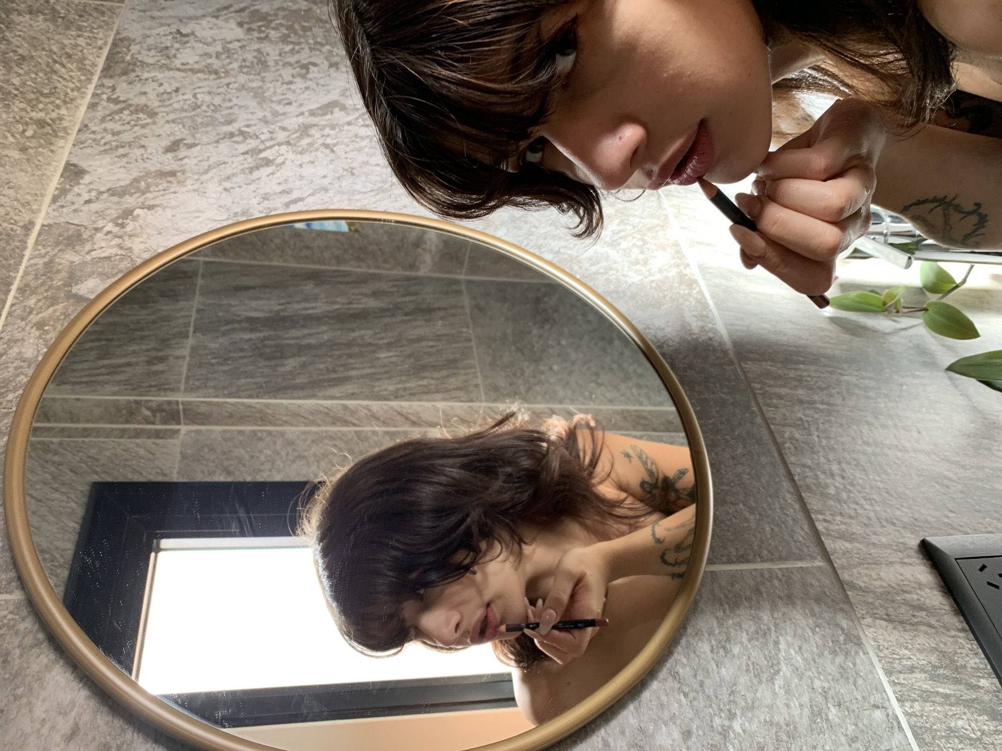A woman with dark brown hair and bangs is applying lip liner while looking in a round gold-framed mirror. The mirror reflects her image against a backdrop of gray-tiled walls. In the mirror's reflection, a window is visible with bright daylight shining through. The woman is holding a dark brown lip liner pencil. Her face and hands are visible, and there are tattoos on her arm. A green plant with leaves is slightly visible to the right, near an electrical outlet. The environment appears to be a bathroom with gray tile walls. The location is not discernible from the image.