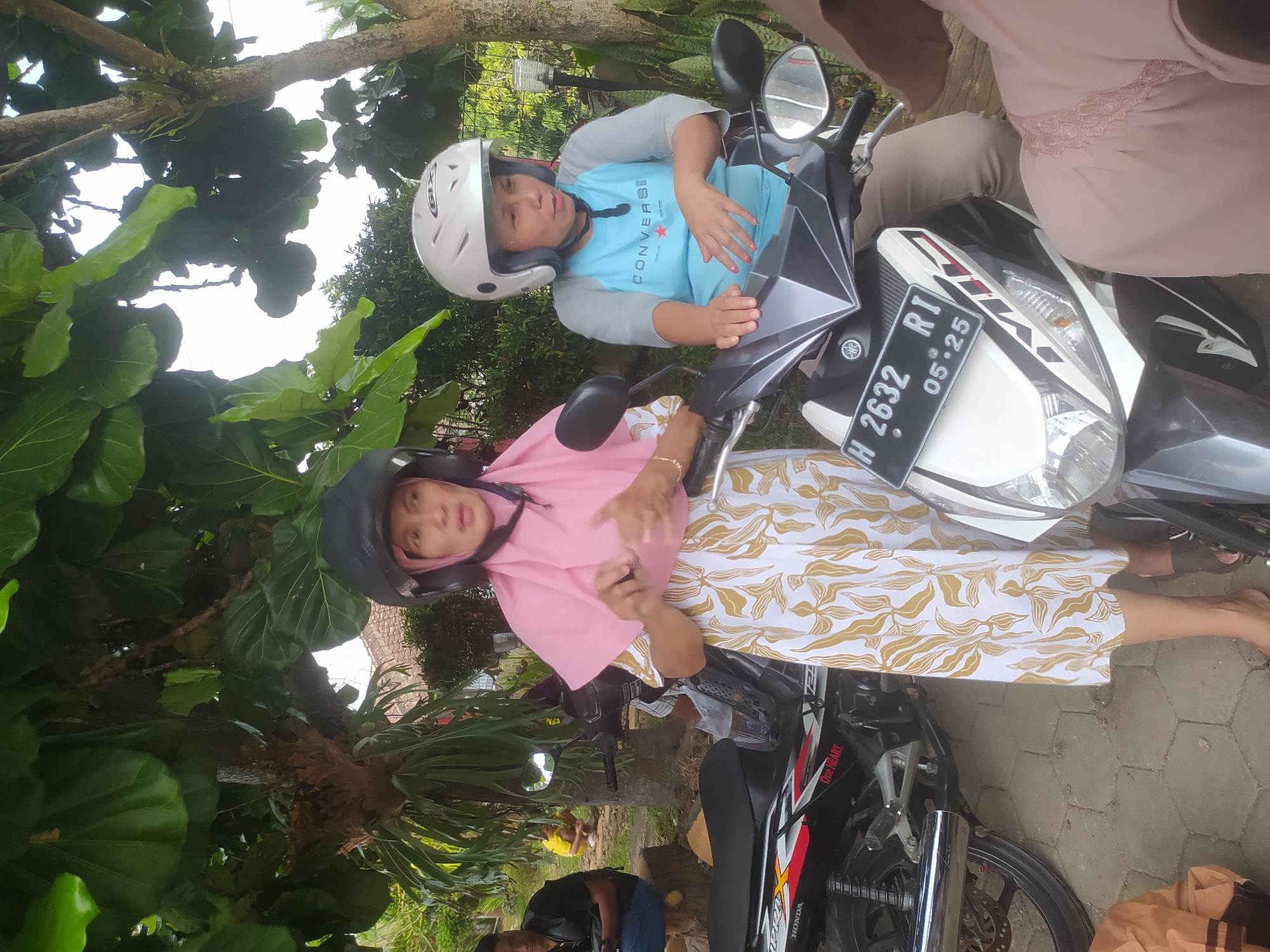 In Candirejo, Indonesia, two adult females, wearing helmets and casual attire, are positioned beside two motorcycles. The female on the left, clad in a pink headscarf and a white and gold patterned dress, stands next to a black Honda motorcycle with red and white accents. The female on the right, wearing a white helmet and a light blue shirt with the "CONVERSE" logo, sits on a white Yamaha motorcycle. The Yamaha bears the license plate "H 2632 RI" and a sticker that reads "05:25". The ground is paved with hexagonal tiles. In the background, a man in a black shirt sits on a wooden log next to a motorcycle. Lush green foliage from a large tree overhangs the scene, and a fence is visible further in the background.
