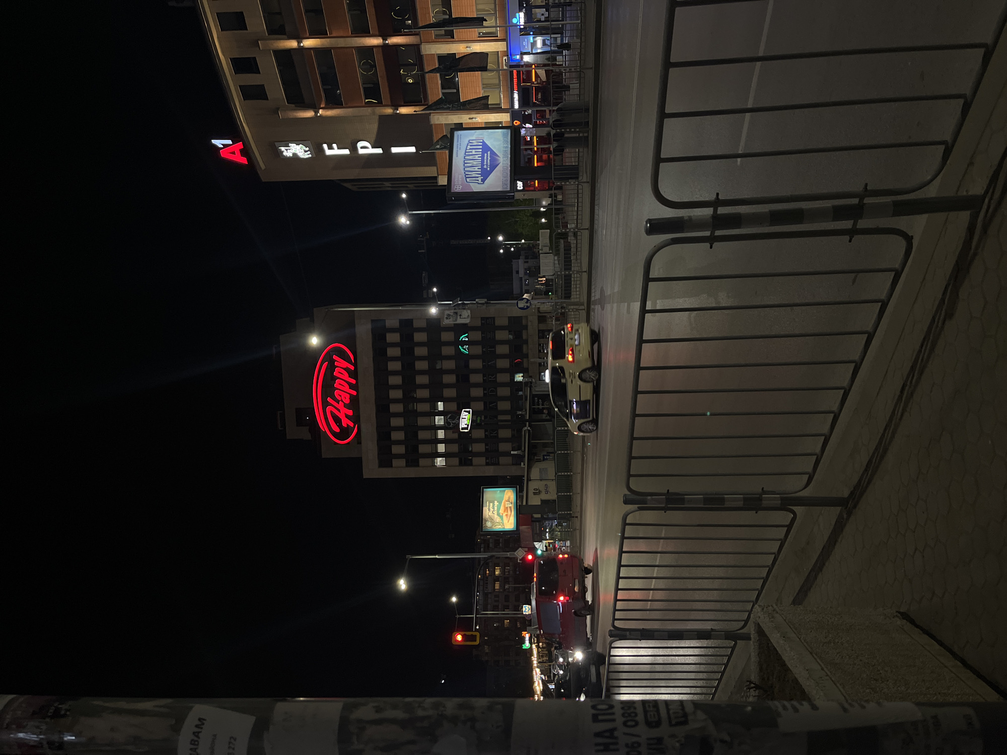 The image, captured at night in Sofia, Bulgaria, presents a vibrant urban street scene. The view looks down a multi-lane road flanked by commercial buildings, with a dark sky overhead. Numerous illuminated signs cast light onto the street and surrounding areas, characteristic of a city after dark.
In the mid-ground, a prominent building features a large, glowing red "Happy" logo, indicating a popular restaurant chain. Above it, on another building, the red "A1" logo for a telecommunications company is visible, alongside white "FPI" and "PULEV" text on other facades. A brightly lit billboard advertises "ДИАМАНТИ" (Diamonds) with additional Cyrillic text reading "ПАЗАРЪТ НАЙ-НОСИ РИСК ОТ РАЗВИВАНЕ НА ЗВО" (The market carries the highest risk of developing zvO). Smaller "SHOP" signs are also present.
The road below shows active traffic, with several cars identifiable by their headlights and taillights. One traffic light displays a red signal, suggesting vehicles are stopped or slowing. Streetlights illuminate the pavement and road. To the right, metal barriers line the sidewalk, separating pedestrians from the traffic lanes. In the immediate foreground on the left, part of a pole or advertising column is visible, adorned with various scraps of paper or graffiti featuring text such as "ABAM", "АРЕНА", "272", "НА ПО", "06/0890", and "BR". The overall impression is one of a bustling city center after dark.