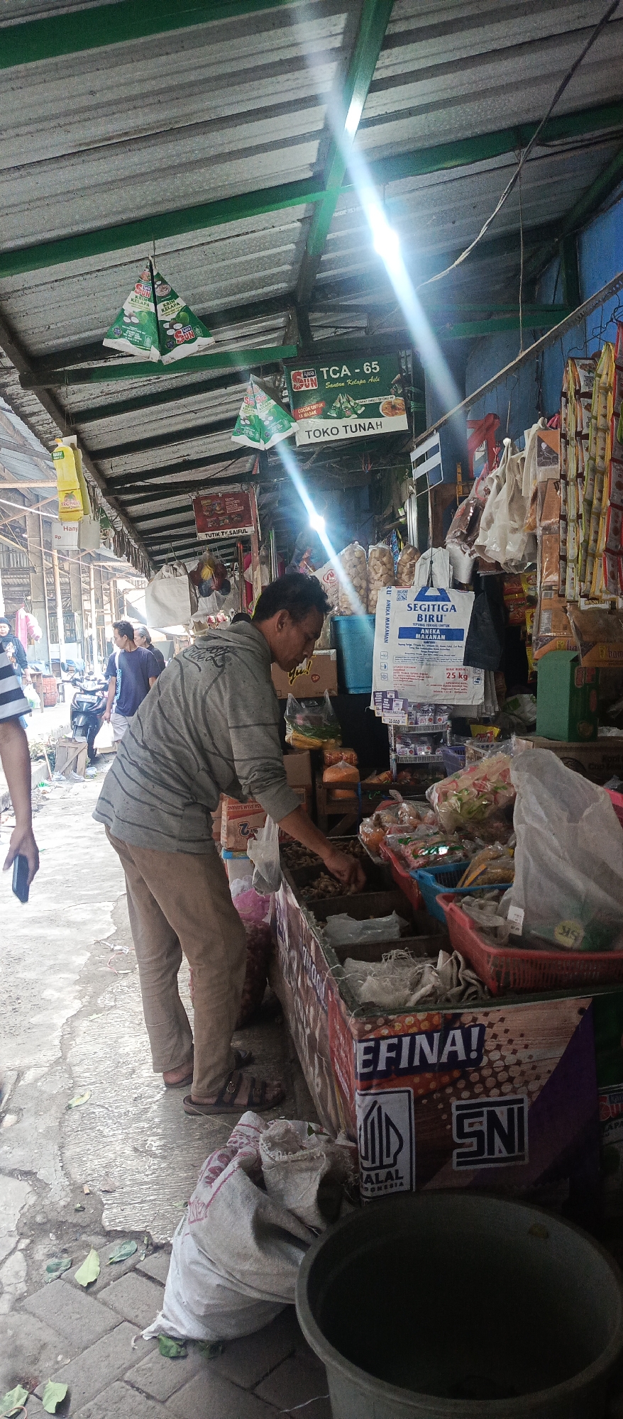 The image depicts an outdoor market stall in Malang, Indonesia. A man in a grey striped shirt and tan pants is browsing through goods displayed in a wooden cart. The cart is filled with various items, including what appear to be dried goods and spices. Behind the cart, a variety of packaged food items and household products are visible, with some hanging from the stall's roof. The stall has a corrugated metal roof supported by green metal beams, suggesting a basic, open-air structure typical of local markets. In the background, other market-goers and stalls can be seen, along with a motorcycle, indicating a busy commercial environment. The ground is paved with concrete and bricks. Sunlight streams in from the upper right, creating a bright lens flare and casting shadows.