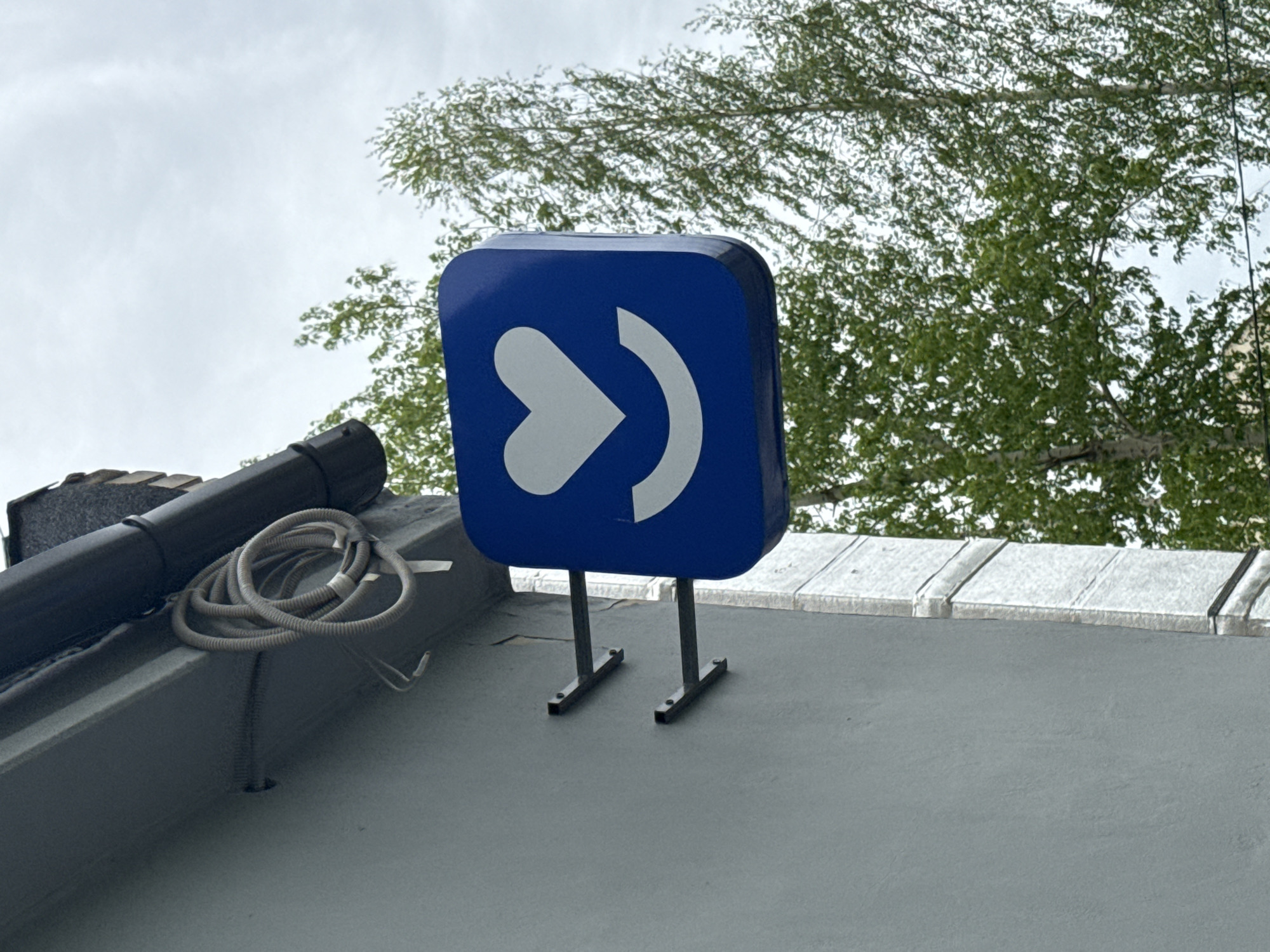 A square blue sign with a white heart-shaped arrow pointing right sits on a roof in an outdoor setting. The sign is mounted on two black metal supports. To the left, a black downspout and coiled gray wiring are visible. In the background, there is a tree with green leaves and a cloudy sky. The location is Unknown, Unknown.