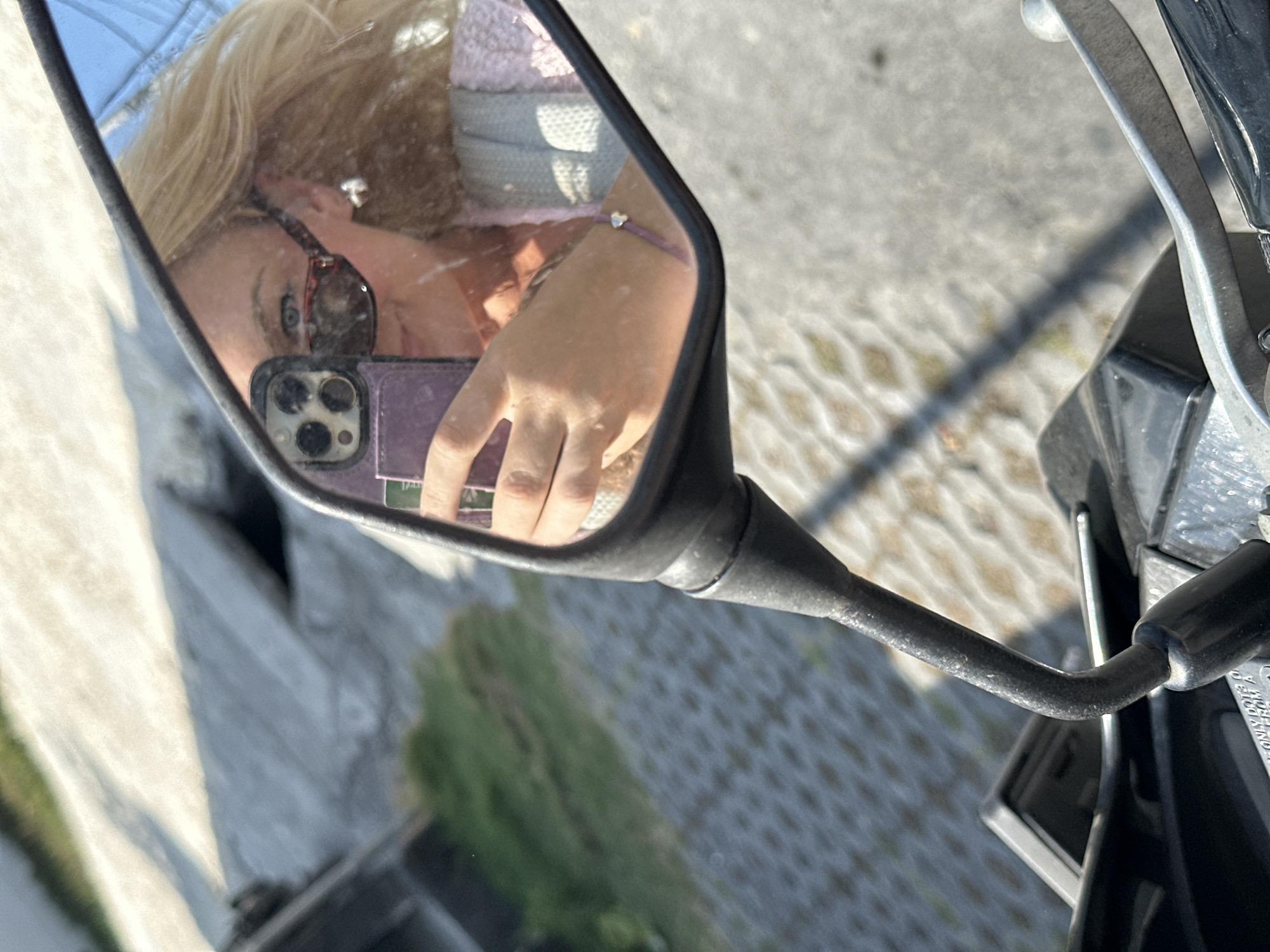 In a motorcycle mirror, a woman with blonde hair and sunglasses is reflected. She is holding a purple cell phone, taking a selfie. She is wearing a light blue and pink knitted sweater and a silver heart-shaped earring. A thin purple bracelet with a silver charm adorns her wrist. The background shows a paved ground with a shadow of a person walking and some green vegetation. The image appears to be taken in the city of North Kuta, Indonesia.