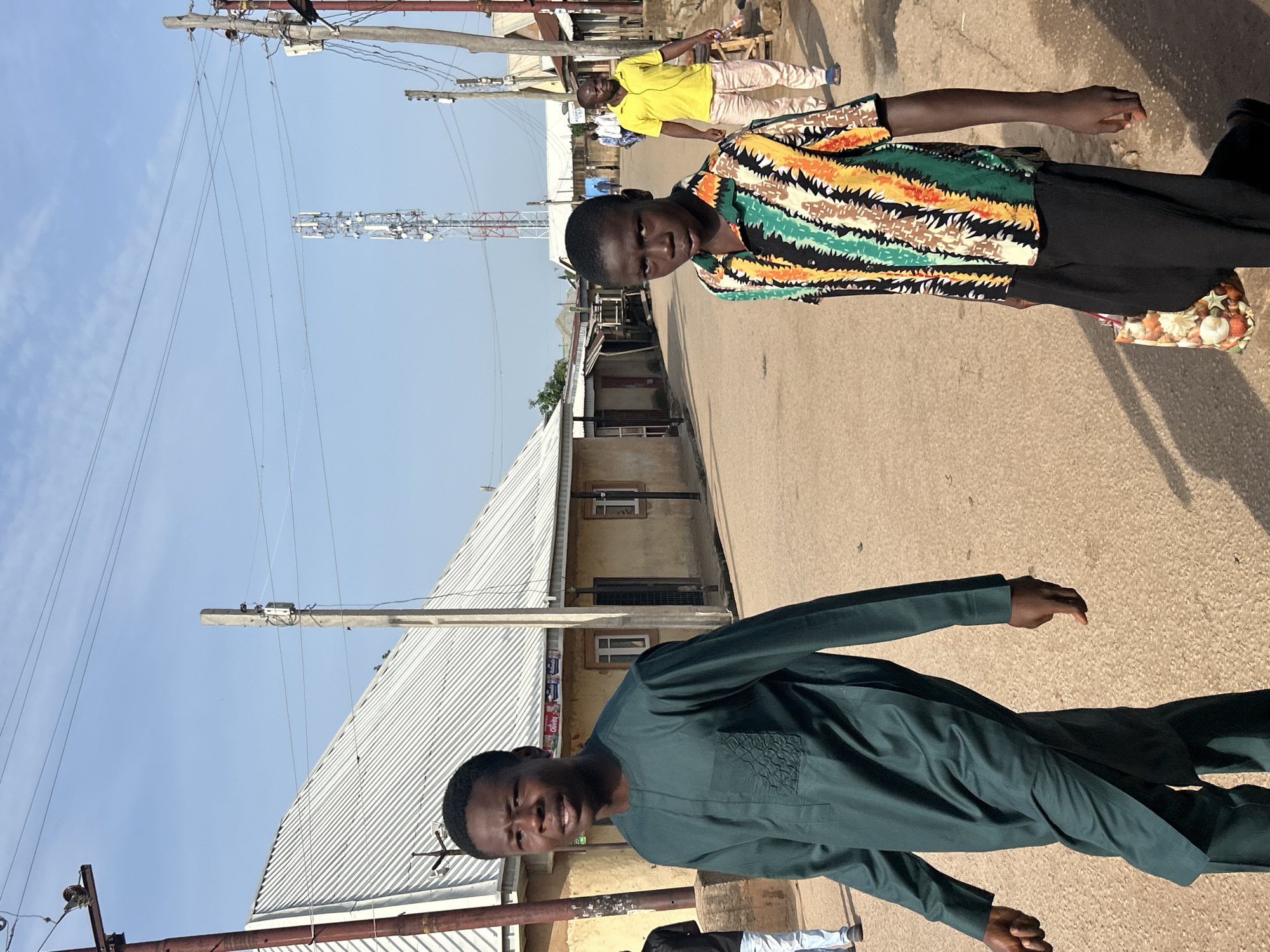 In Jos, Nigeria, a street scene unfolds under a clear blue sky. Two young men walk towards the camera on a dusty road lined with buildings and utility poles. The man on the left wears a dark green, long-sleeved traditional outfit. The man on the right is dressed in a colorful, short-sleeved patterned shirt and black trousers, carrying a patterned bag in his left hand. Behind them, a man in a yellow t-shirt and light-colored trousers walks away from the camera, and other individuals are visible further down the street. A prominent cell tower stands in the background, indicating modern infrastructure within the urban environment. The buildings appear to be single-story structures with corrugated metal roofs. The overall atmosphere suggests a typical day in a Nigerian city.