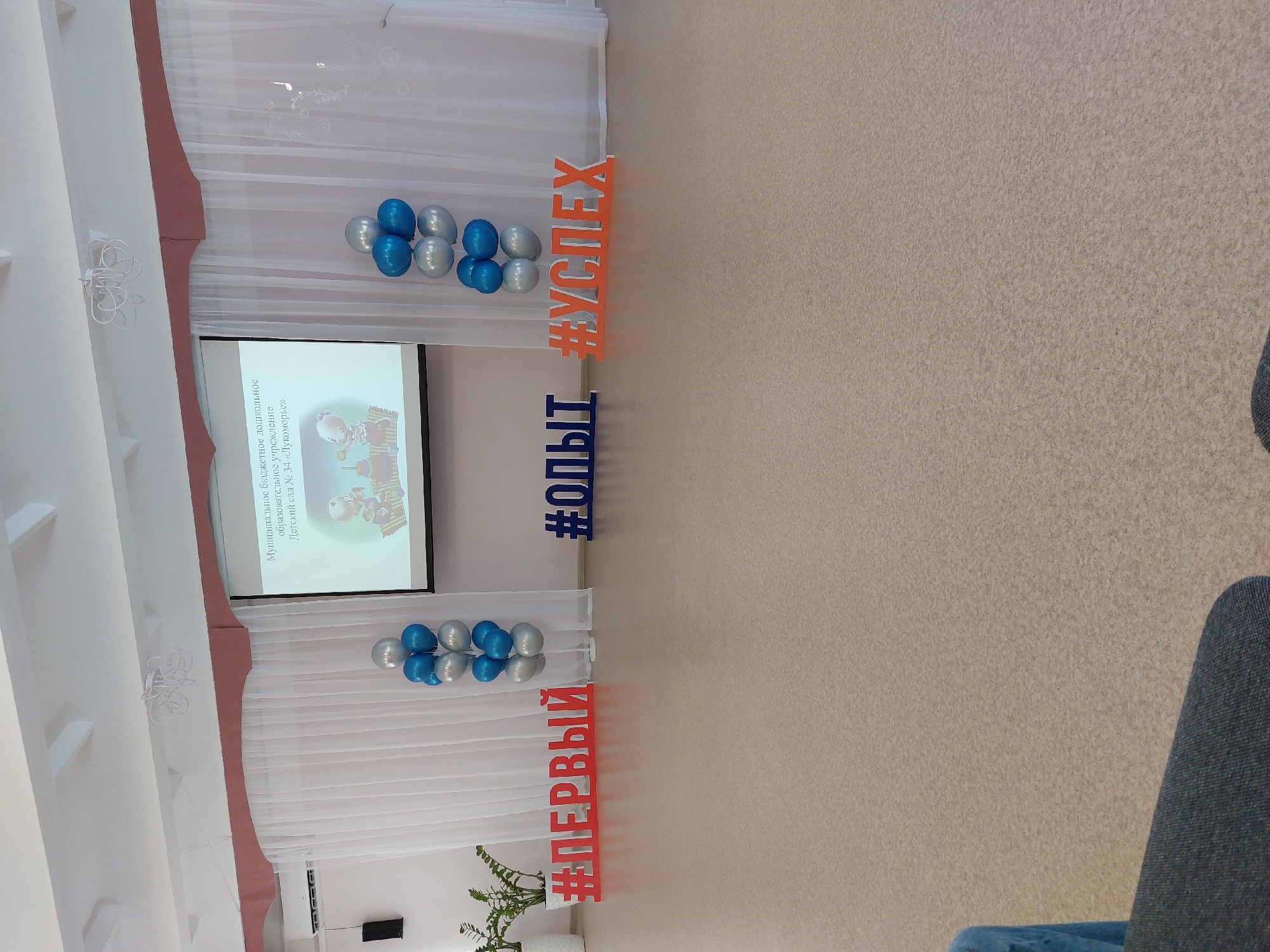 Here's a description of the image:
The image shows a room, likely a kindergarten or preschool, decorated for an event. In the background, there is a white curtain with pink accents hanging behind a projection screen that is showing a presentation slide. The screen displays Russian text, including the name of the institution "Детский сад № 34 «Лукоморье»", which translates to "Kindergarten No. 34 'Lukomorye'". The location is likely Chaikovsky, Russia, based on the name "Chaikovsky" in the location settings.
Arranged around the screen are sets of blue and silver balloons. Below the screen are large, freestanding letters spelling out words in Russian, with hashtags in front of them. The words read "#ПЕРВЫЙ" (First), "#ОПЫТ" (Experience), and "#УСПЕХ" (Success). A potted plant is visible near the bottom left corner of the image. The foreground of the image is mostly a light beige carpet. A piece of furniture, probably a chair, is visible in the bottom right corner. The room appears well-lit.