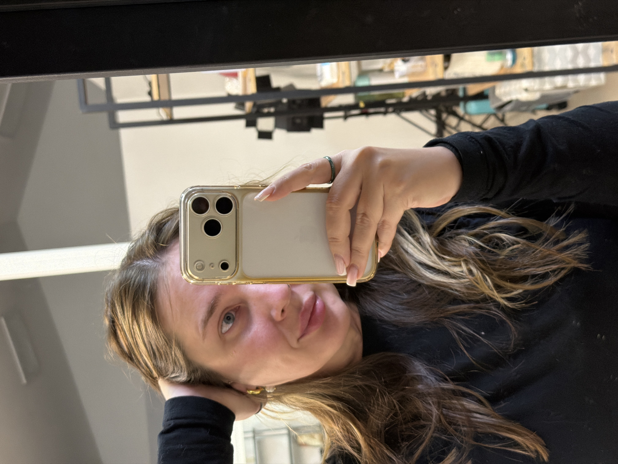 A woman is taking a selfie in front of a mirror. She is holding a smartphone in her right hand, obscuring part of her face. She has light skin, blue eyes, and long brown and blonde hair. She is wearing a black long-sleeved shirt and an earring is visible on her left ear. A ring is visible on her finger.
The mirror reflects a cluttered room with various items on a table or shelf. A black frame surrounds the mirror. The location in the city of Chaikovsky, Russia, cannot be determined from the provided visual information.