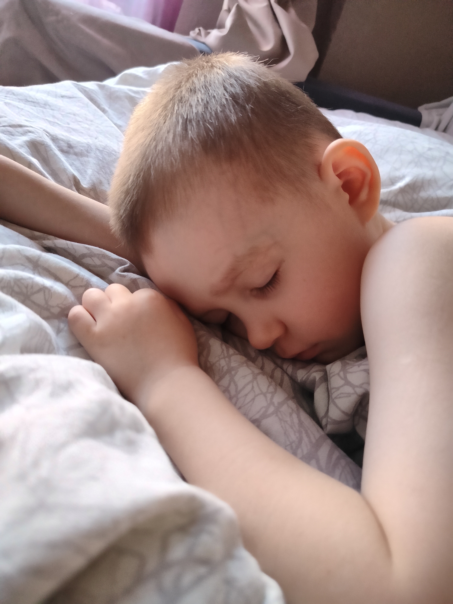 The image captures a young child sleeping soundly on a bed. The child, with light brown hair and fair skin, is lying on their right side with eyes closed, their face gently pressed into a patterned pillow or sheet. One hand is tucked near their head, while the arm and shoulder are visible, resting under or partially covered by light-colored bedding. The bed is covered with a light grey sheet featuring a subtle, geometric pattern of interlocking lines or branches, and a plain light-colored duvet or blanket is also visible. The lighting is soft and natural, indicating daytime, possibly morning or early afternoon, which contributes to the peaceful and serene atmosphere of the scene. The setting appears to be a bedroom. This tranquil moment takes place in Chaikovsky, Russia. There is no visible text in the image.