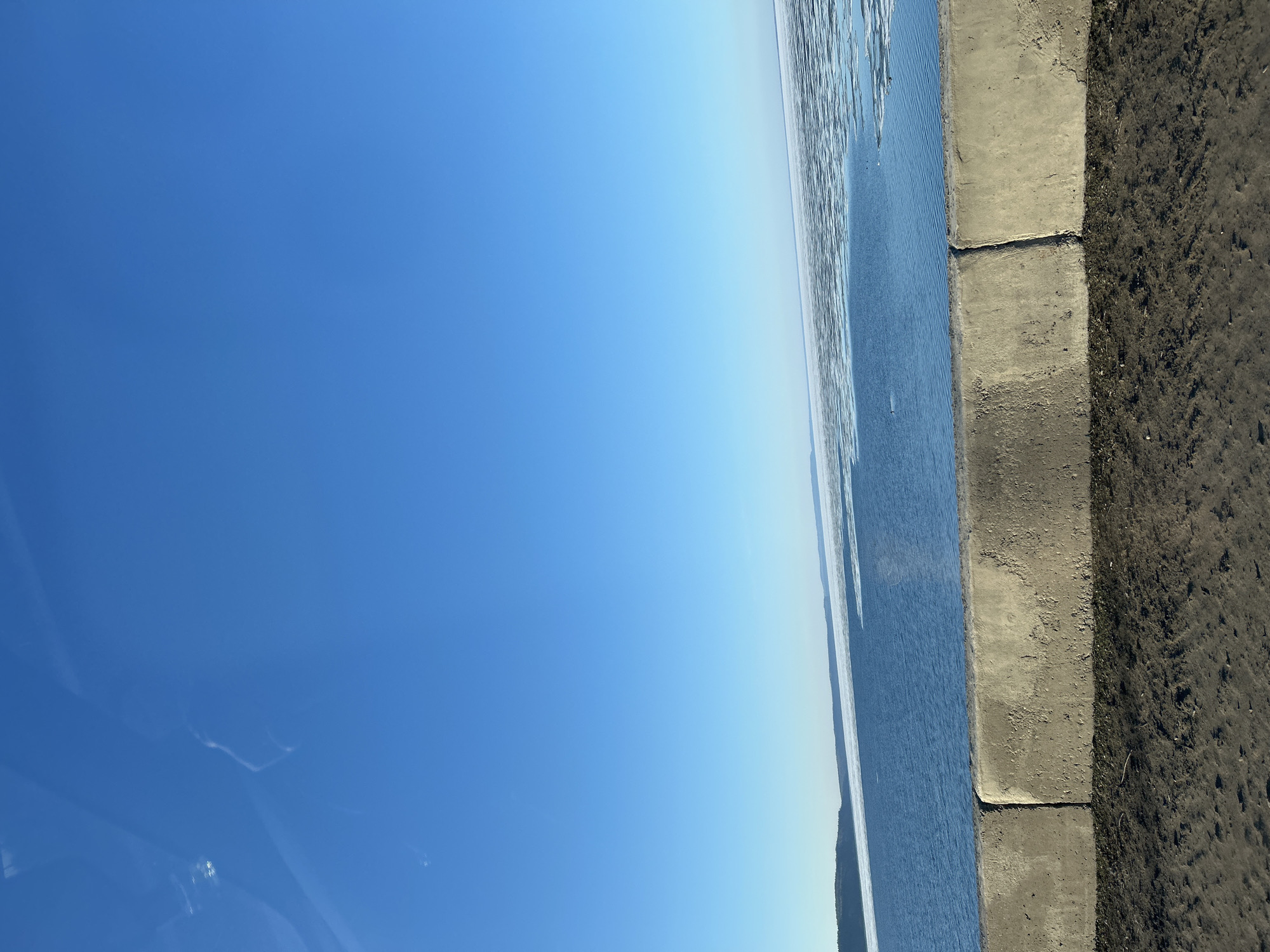 The image shows a clear blue sky above a body of water with ice formations along the edge. A concrete embankment separates the water from a dirt area. It seems to be a cold sunny day in Chaikovsky, Russia, near a body of water such as the Kama River.