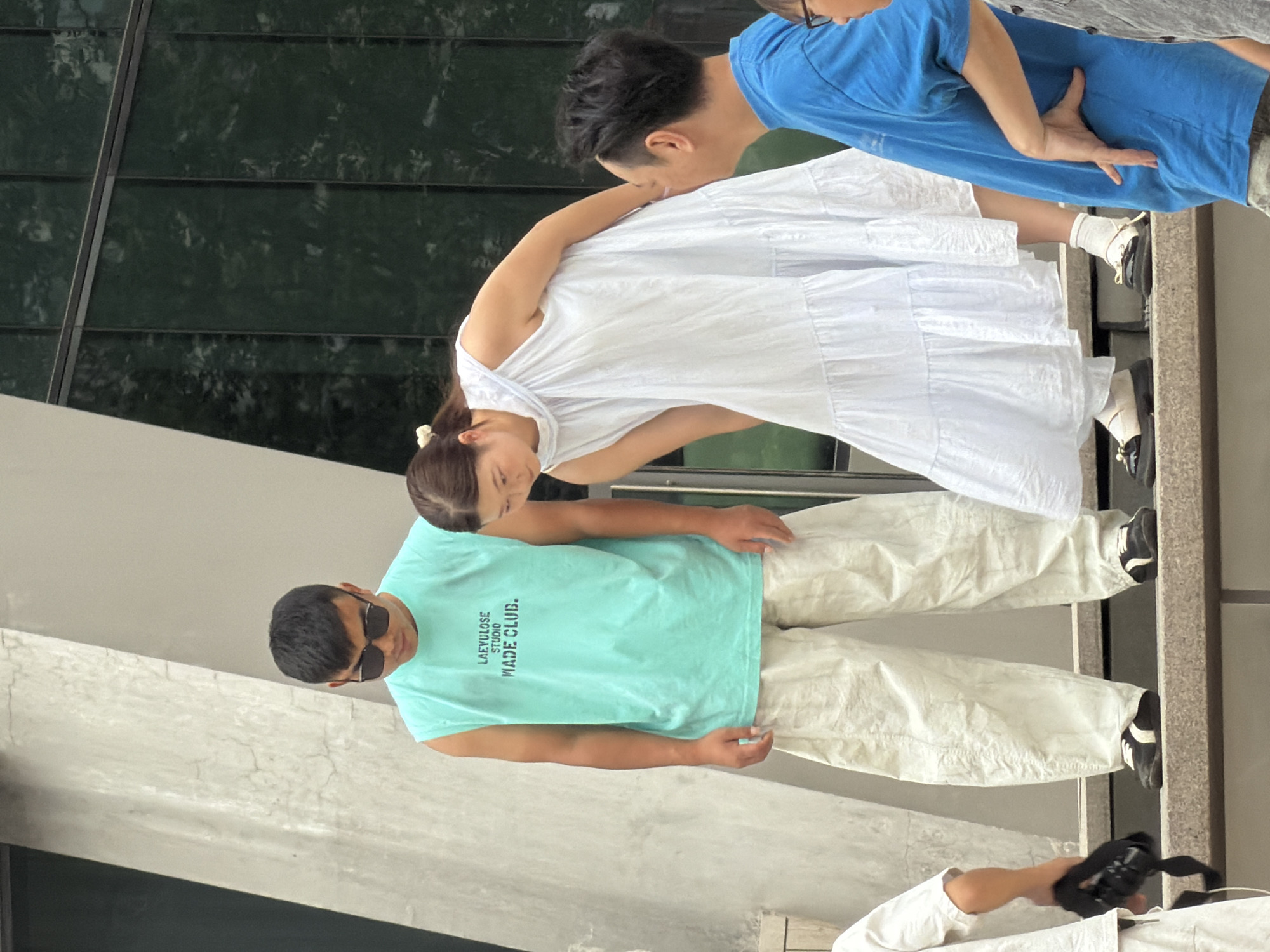 In Yuzhong District, China, three individuals are positioned on outdoor steps in front of a building with a dark, reflective glass facade. On the left, a man wearing sunglasses and a teal sleeveless t-shirt with "LAEVULOSE STUDIO MADE CLUB." printed on it, stands facing downwards. He is dressed in loose, cream-colored pants and black and white sneakers. To his right, a woman in a flowing white sleeveless dress with tiered ruffles and white socks, leans forward, looking downwards and away from the camera. She is wearing black and cream flat shoes. To the right of the woman, a man in a bright blue t-shirt is facing the woman, his right hand resting on his hip and his left hand partially visible. He is wearing glasses. Partially visible on the far left is a person in a white short-sleeved shirt, holding a black camera with a strap. The steps appear to be made of concrete or a similar material.