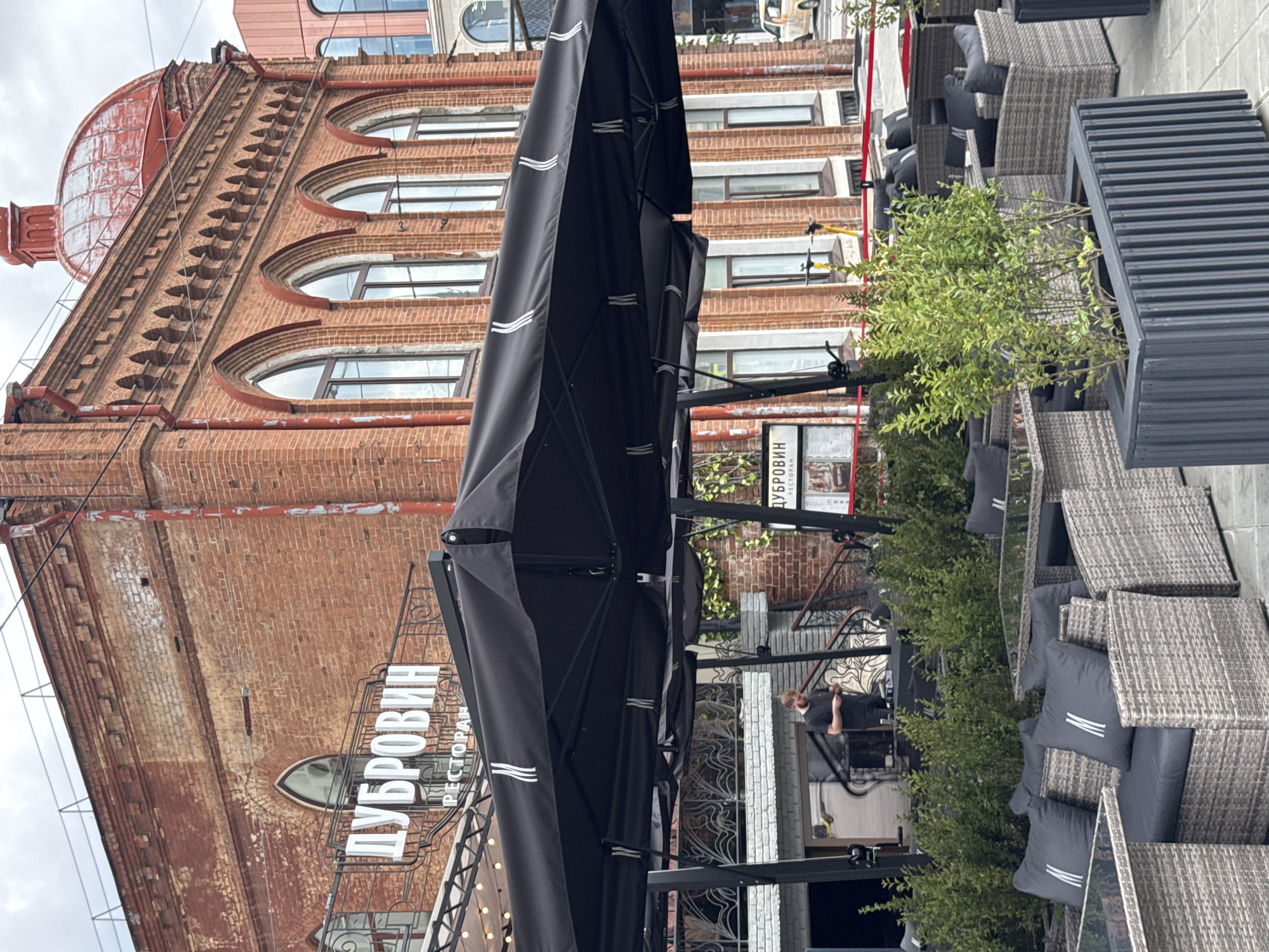 The image depicts the exterior of a restaurant named "ДУБРОВИН" (Dubrovin) located in Yekaterinburg, Russia. The restaurant is housed in a historic brick building with arched windows and a red domed roof. In front of the building, there is an outdoor seating area with tables and chairs covered by large black umbrellas. The seating consists of wicker furniture with dark gray cushions, some of which have a white "M" logo. A man in a black t-shirt is standing near the entrance, looking at his phone. Several potted plants add greenery to the outdoor space. The scene appears to be a casual outdoor dining setting on an overcast day.