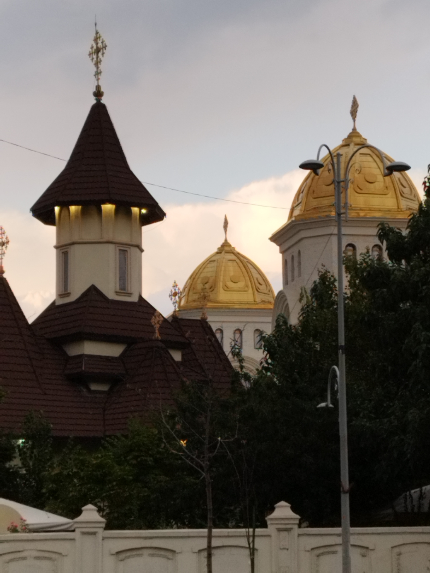 The image shows the exterior of a religious building, likely a church or cathedral, in Bucharest, Romania. The building features multiple distinct architectural elements including several steep, dark-tiled roofs, a tall, light-colored tower topped with a cross, and at least two prominent golden domes. The sky is overcast, suggesting either dawn, dusk, or a cloudy day. Lush green trees partially obscure the lower portions of the building and extend into the foreground. In front of the building, a white decorative wall with arched sections is visible, further suggesting an urban setting. A street light is positioned in the foreground, to the right of the center, adding to the urban context. The image appears to be taken from ground level, looking up at the structure.