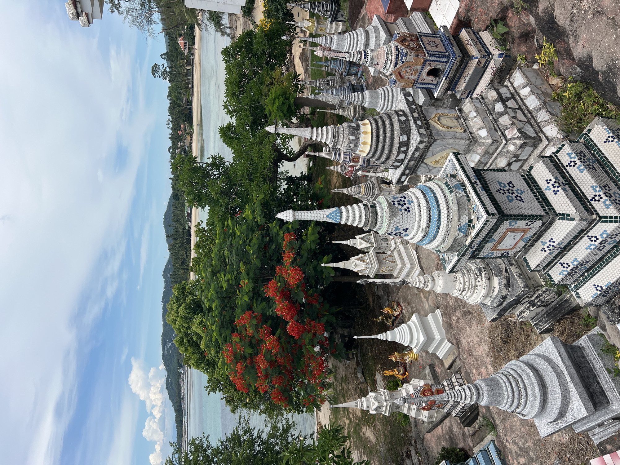 The image depicts a tiered arrangement of small, ornate stupa-like structures and statues, likely from a temple or memorial site, overlooking a tropical coastline. The foreground and midground are dominated by these structures, which vary in size, material, and decorative detail, with many featuring intricate mosaic work and spire-like tops. Some gold-colored statues of deities are visible amongst the stupas. Behind the structures, lush green trees are abundant, with one tree prominently displaying vibrant red flowers. The background reveals a calm body of turquoise water meeting a sandy beach, backed by a low, verdant hill dotted with buildings. The sky is partly cloudy with a bright blue expanse. The scene is likely located in Baan Hua Thanon, Thailand, given the architectural style and tropical environment. There are no people visible in the image.
