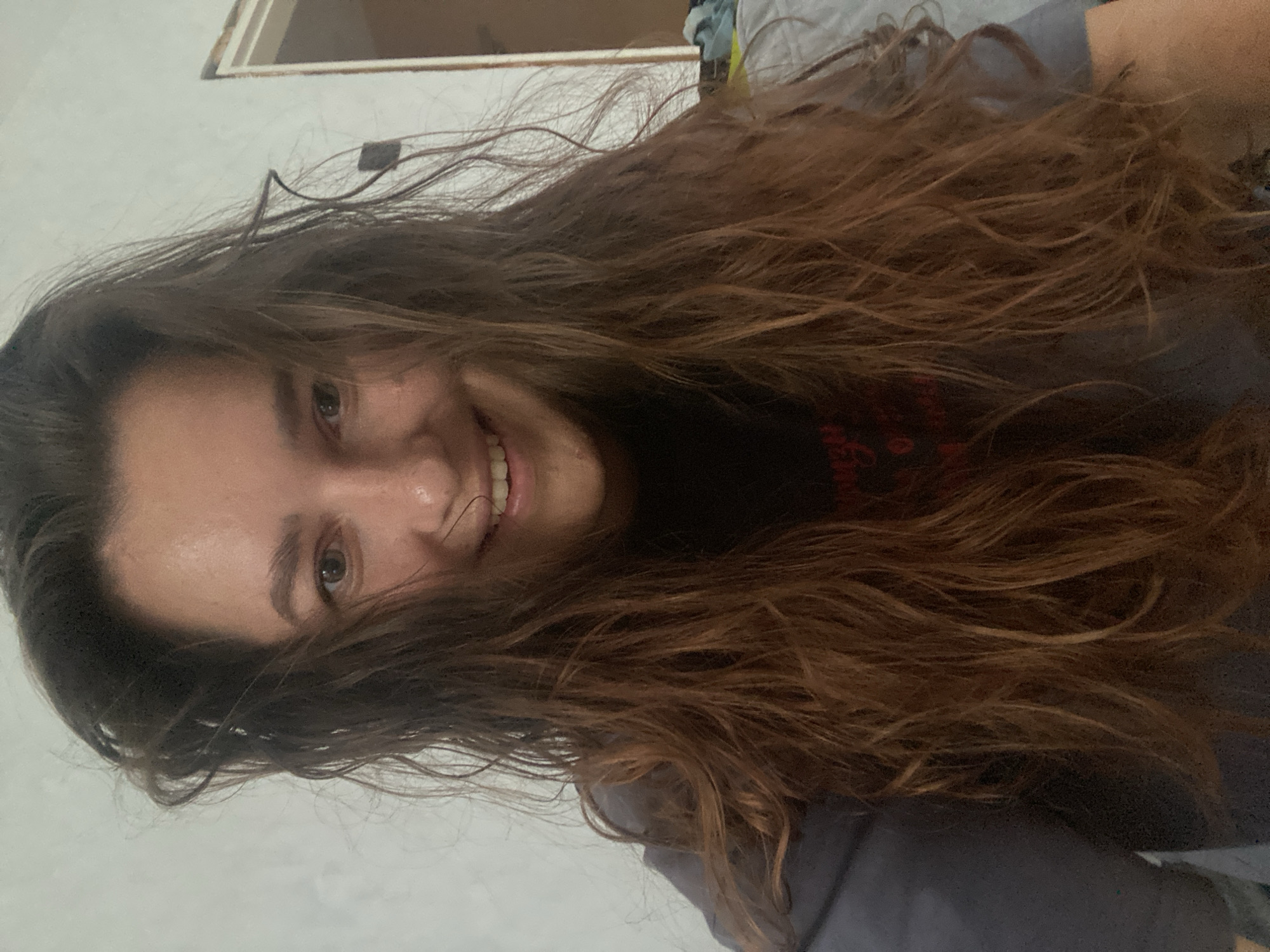 The image shows a close-up, tilted shot of a young woman's face and long, dark brown, wavy hair. She has fair skin, brown eyes, and a wide, genuine smile with her teeth showing. Her hair is somewhat messy and extends across the frame, partially obscuring her face. She is wearing a dark-colored t-shirt. The background appears to be indoors, with a white wall visible on the left and a hint of what might be a shelf or furniture in the upper right. The lighting suggests it could be daytime. The image evokes a casual and happy mood. There is no visible text.