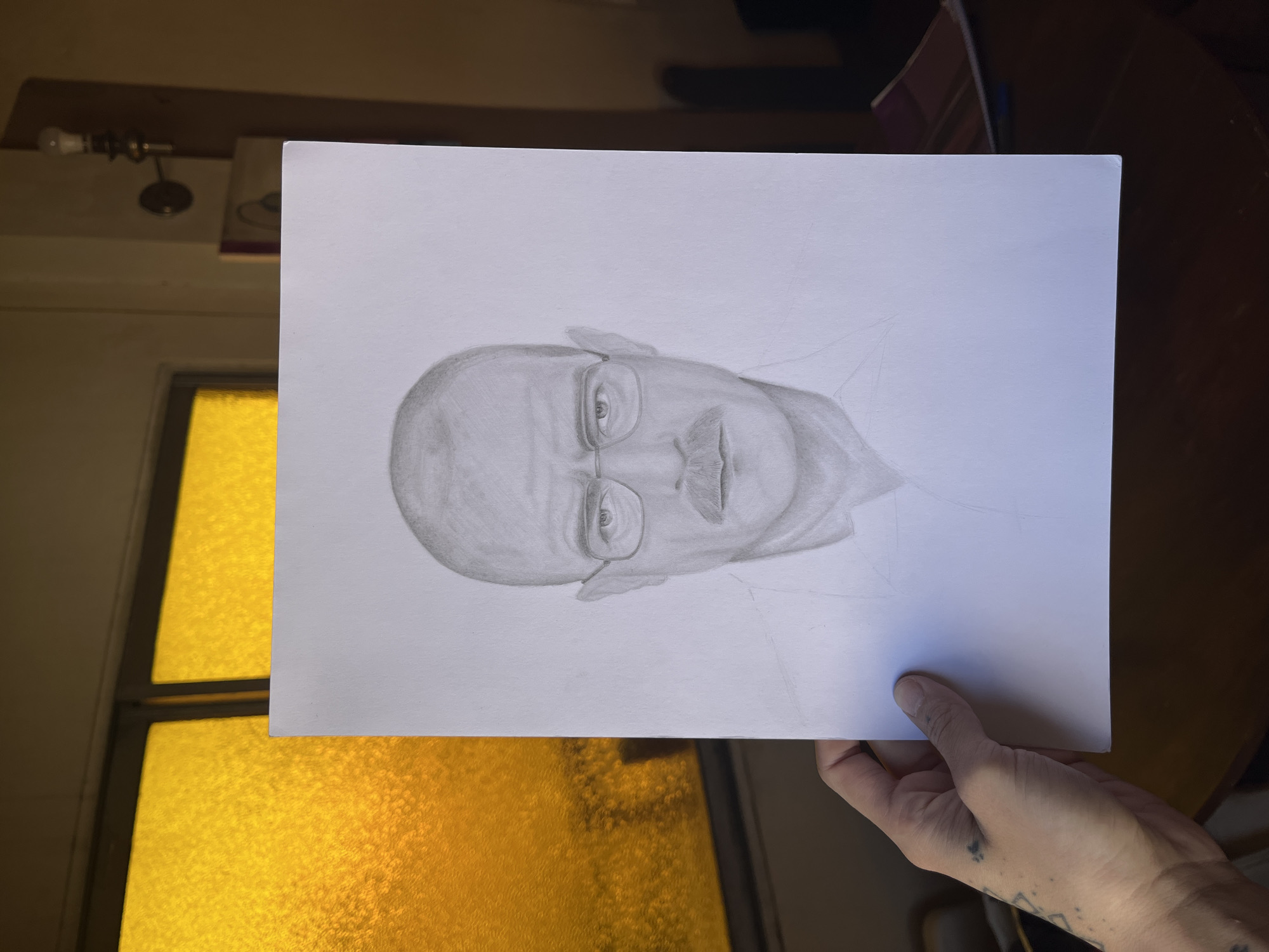 An image, rotated clockwise, displays a white sheet of paper holding a pencil drawing, held vertically by a person's left hand. The drawing is a realistic, monochromatic portrait of a bald man with glasses and a prominent mustache, depicting his head and upper chest, notably resembling the character Walter White. The hand holding the paper features several small, dark tattoos on the wrist and lower forearm.
The setting is an indoor space, possibly a room in Buenos Aires, Argentina. In the background, a window with highly textured, yellow-orange glass casts a warm, vibrant glow. Above the window, a bare light bulb in a brass-colored fixture is mounted on a wooden beam or shelf. Partially visible in the upper right background are dark objects, likely books or files, alongside a blue pen.
The activity is the presentation or photography of the drawing. Notable details include the preliminary sketch lines visible on the right side of the drawing, suggesting it is unfinished. The warm hue from the window implies either a specific type of colored glass or a late time of day. No visible text is present in the image.