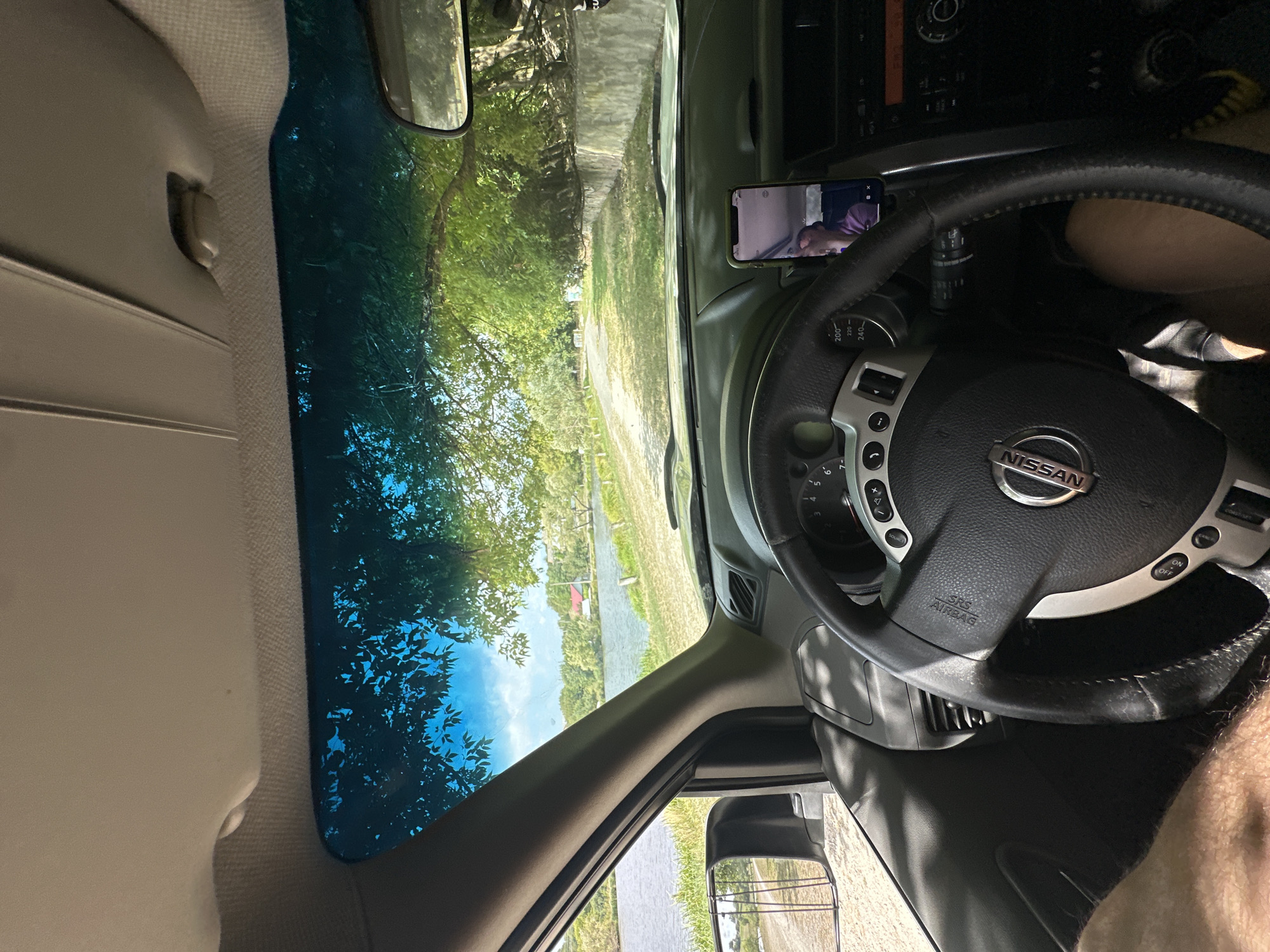 The image shows the interior of a car, viewed from the driver's perspective. The dashboard, steering wheel, and part of the driver's legs are visible. Outside the car, a body of water, a dirt road, and trees can be seen. The scene appears to be a rural or park-like setting. The location is identified as Makiv, Ukraine.