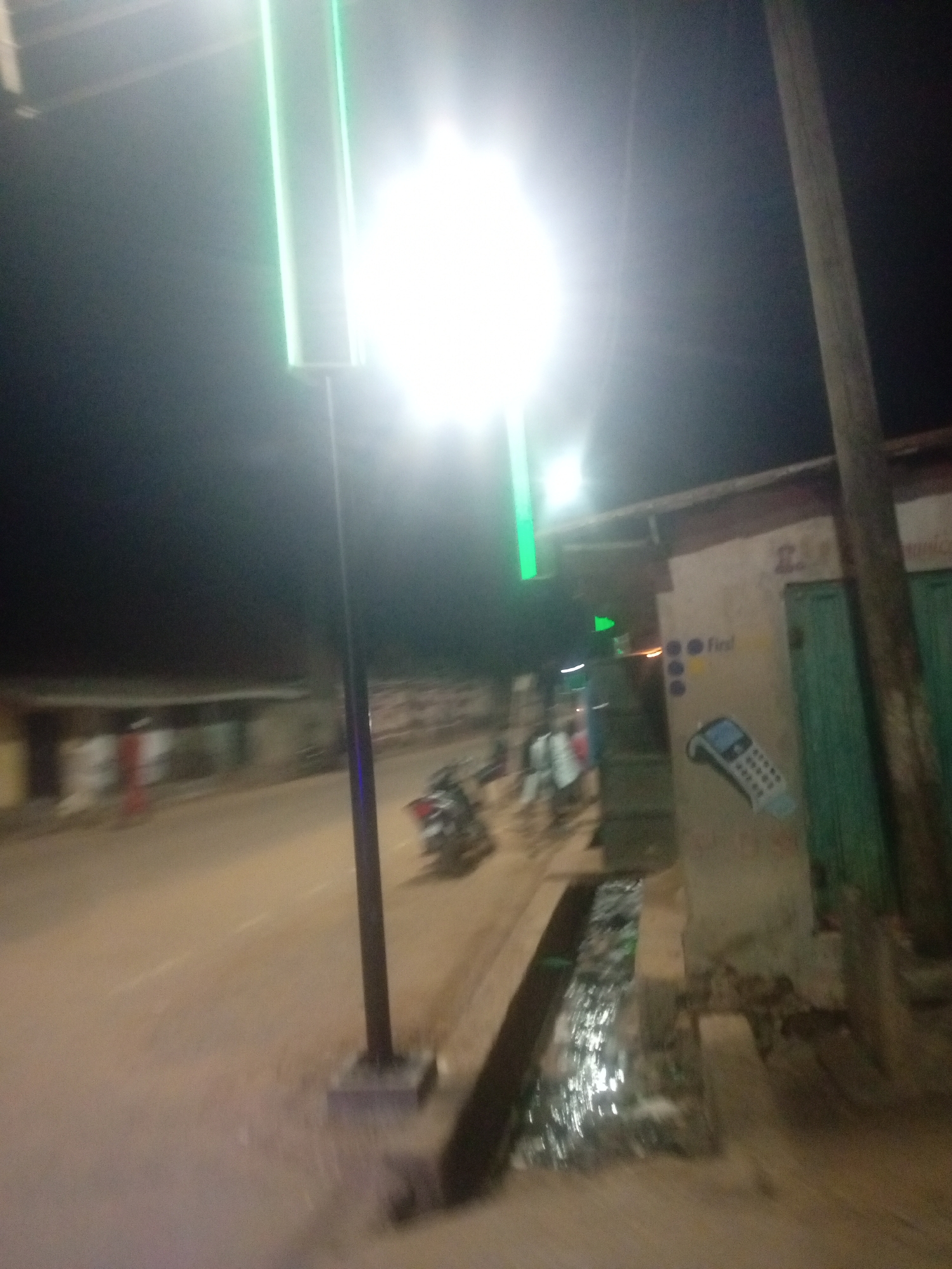 The image depicts a street scene at night in Tundun Wada West, Nigeria. A tall, lit signpost with bright green vertical lights stands prominently on the left side of the road, illuminating the surrounding area with an intense glow. The road itself appears to be unpaved and slightly damp, with a visible drainage channel running alongside it.
To the right of the road, a building with a light-colored wall is visible. On this wall, there are several markings, including what appears to be a graphic of a mobile phone or a card reader. Above the graphic, there are three blue circles and the word "first" written in yellow. A green door is also visible on this building.
Further down the street, on the left side, blurred figures and a motorcycle are discernible, suggesting some activity and movement. The overall atmosphere is dark, characteristic of nighttime, with the bright artificial lights providing the primary illumination. The scene suggests a commercial or residential area in a developing locality, with the street serving as a central thoroughfare. No specific weather conditions are evident, but the dampness of the road might imply recent rain. The image conveys a sense of everyday life occurring at night in this Nigerian location.