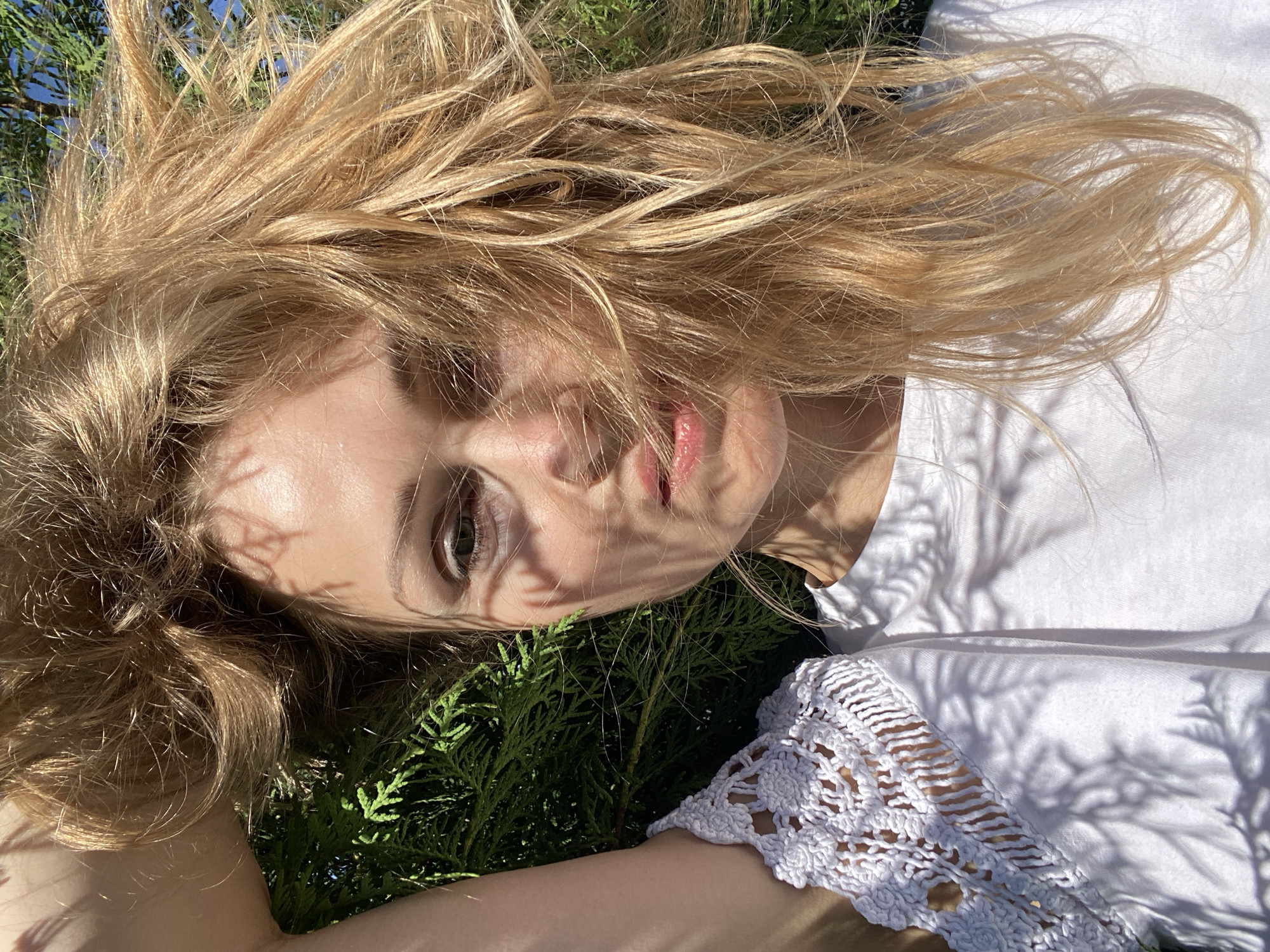 A young Caucasian woman with blonde hair lies on a backdrop of green foliage, possibly in a garden or natural area. Her hair is spread out around her head, and sunlight filters through, casting dappled shadows on her face and white t-shirt. The t-shirt features a white crochet detail on the shoulder. The woman's right arm is raised and her hand is in her hair. Her face is looking towards the camera, with her lips slightly parted. The background shows green leaves and branches, and in the upper right corner, a hint of blue sky and evergreen branches are visible. The overall lighting suggests it is daytime, with bright sunlight. The location is not specified in the image.