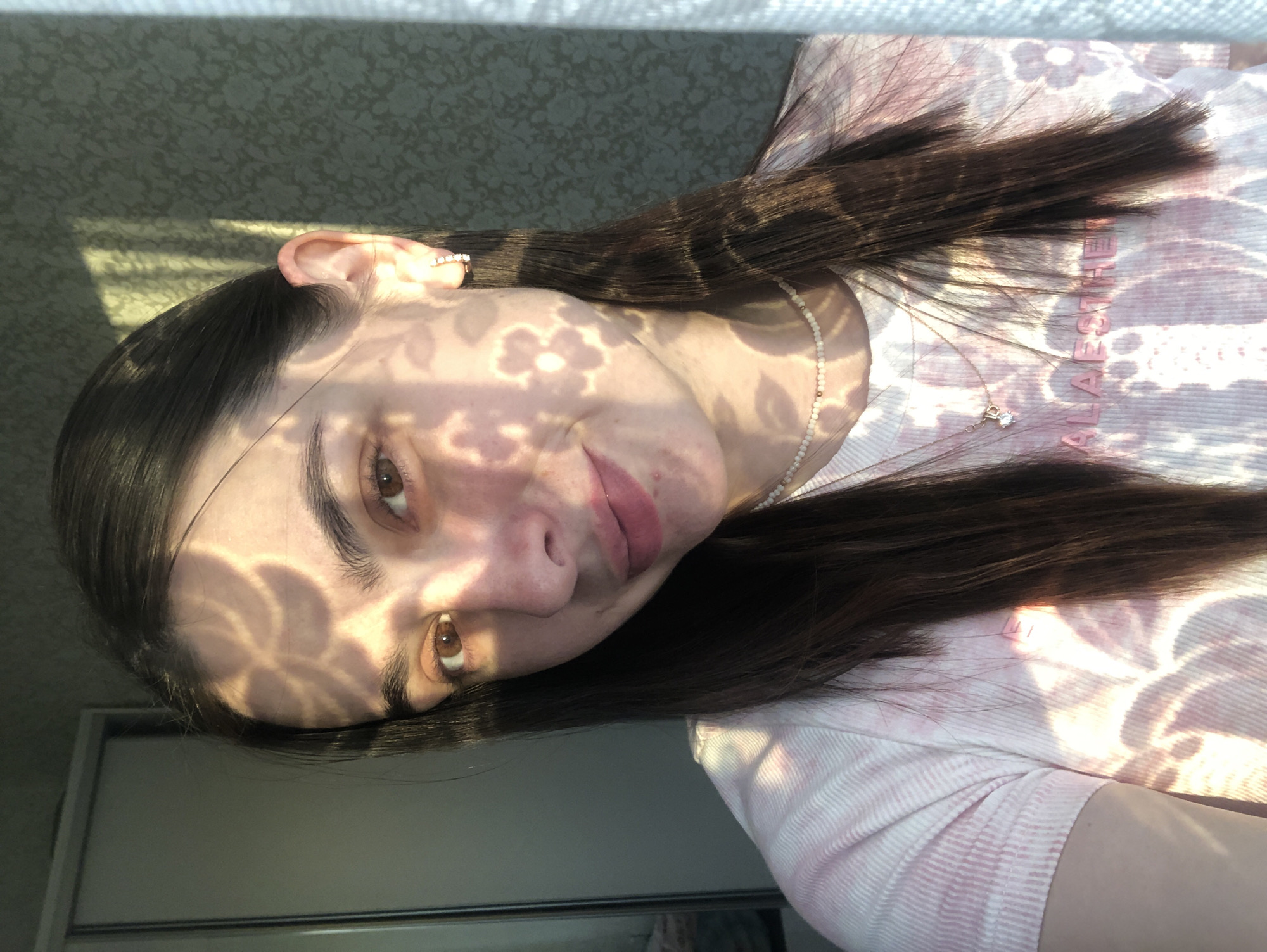 The image is a medium shot of a young woman from the chest up. She has long, dark brown hair and brown eyes. She is wearing a light-colored t-shirt with a pink tie-dye pattern and floral designs. She is also wearing a delicate necklace with small beads and a pendant. A flower-shaped shadow is cast across her face and chest, suggesting the presence of a patterned window or screen. Her skin has some blemishes. She is looking towards the camera with a slight smile. The background shows a wall with a textured pattern, a closet door, and a window with a white curtain. The lighting suggests it is daytime, possibly late afternoon, with sunlight creating the shadows. The woman is likely indoors. There is no clear indication of the specific location within Noviy, Russia.