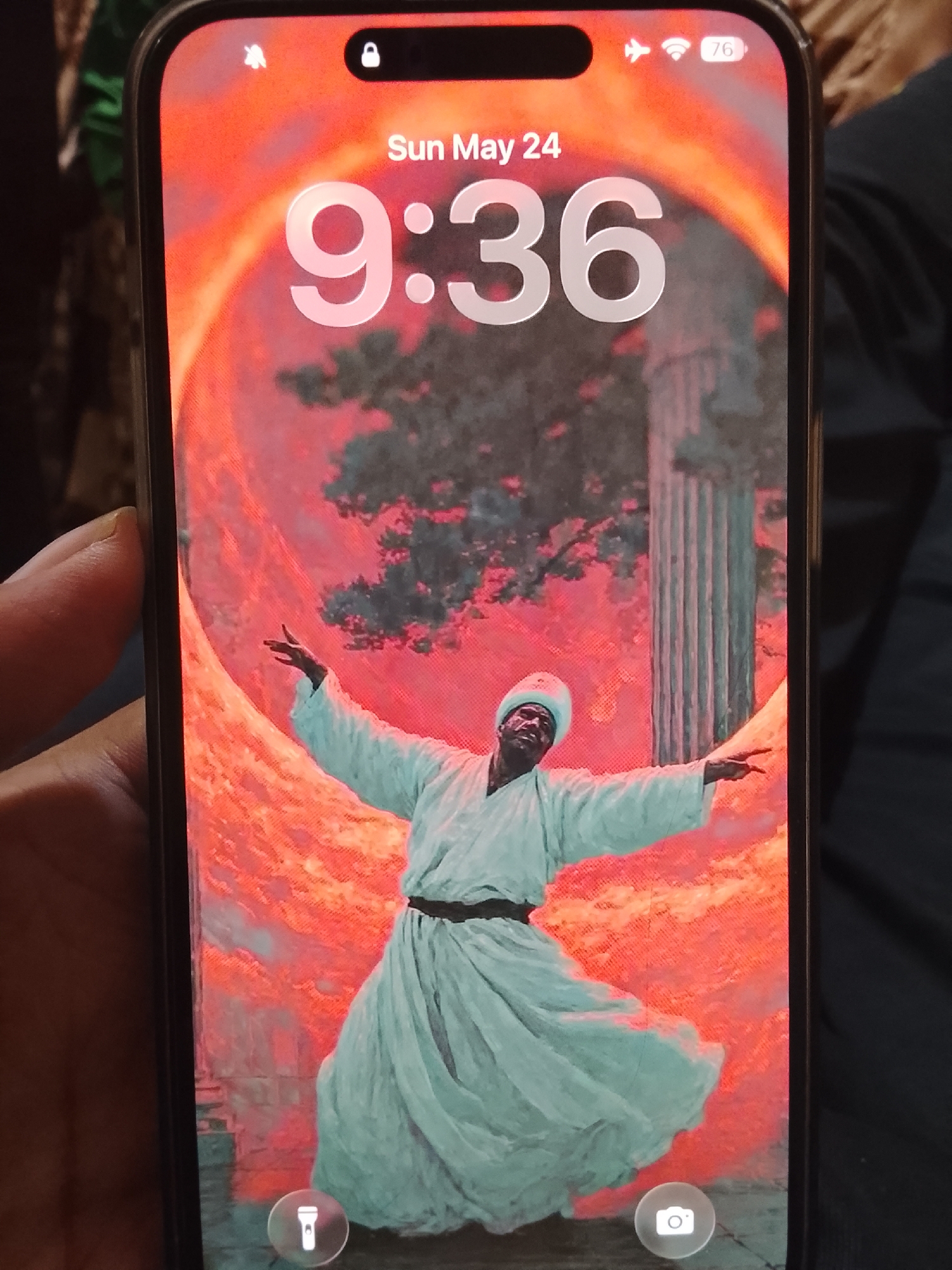 A close-up photo shows an iPhone displaying its lock screen. The screen shows the date "Sun May 24" at the top, with the time "9:36" prominently displayed. The wallpaper features an artistic depiction of a Sufi whirling dervish figure wearing a white robe and a tall, light blue hat. He is set against a background with an orange sun-like orb, foliage, and a classic column. There are two icons at the bottom of the screen: a flashlight icon on the lower left and a camera icon on the lower right. The phone's battery level is at 76%. The image does not contain enough details to connect the background to the city of Multan, Pakistan, as the scene is an artistic rendering rather than a realistic depiction of a location.