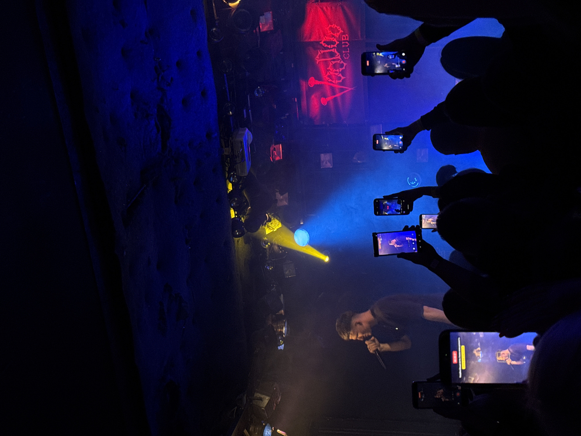 The image depicts a live music performance taking place at night in a dark club, viewed from the audience's perspective. The scene is dominated by a male performer on stage, seen in profile, actively singing into a microphone. He has short dark hair and is wearing a dark t-shirt.
The stage is illuminated by dramatic lighting, featuring prominent yellow and blue spotlights. Behind the performer, a dark banner with stylized red text clearly reads "VOODOO CLUB," identifying the venue. Various stage equipment, including other lights and what appear to be speakers, are dimly visible around the stage area.
In the foreground and mid-ground, several audience members are visible as dark silhouettes. Their hands are raised, holding up smartphones to record or photograph the performance. The screens of these phones are visible and show a clear, real-time capture of the performer on stage. The dark outlines of their heads and shoulders suggest a crowded and engaged audience.
The overall atmosphere is typical of a concert, with the vibrant stage lights creating a focal point against the dark backdrop of the club. The visible text "VOODOO CLUB" serves as a specific location cue for the venue, which is in Warsaw, Poland, as per the provided context. The image captures the energetic and interactive nature of a live music event.