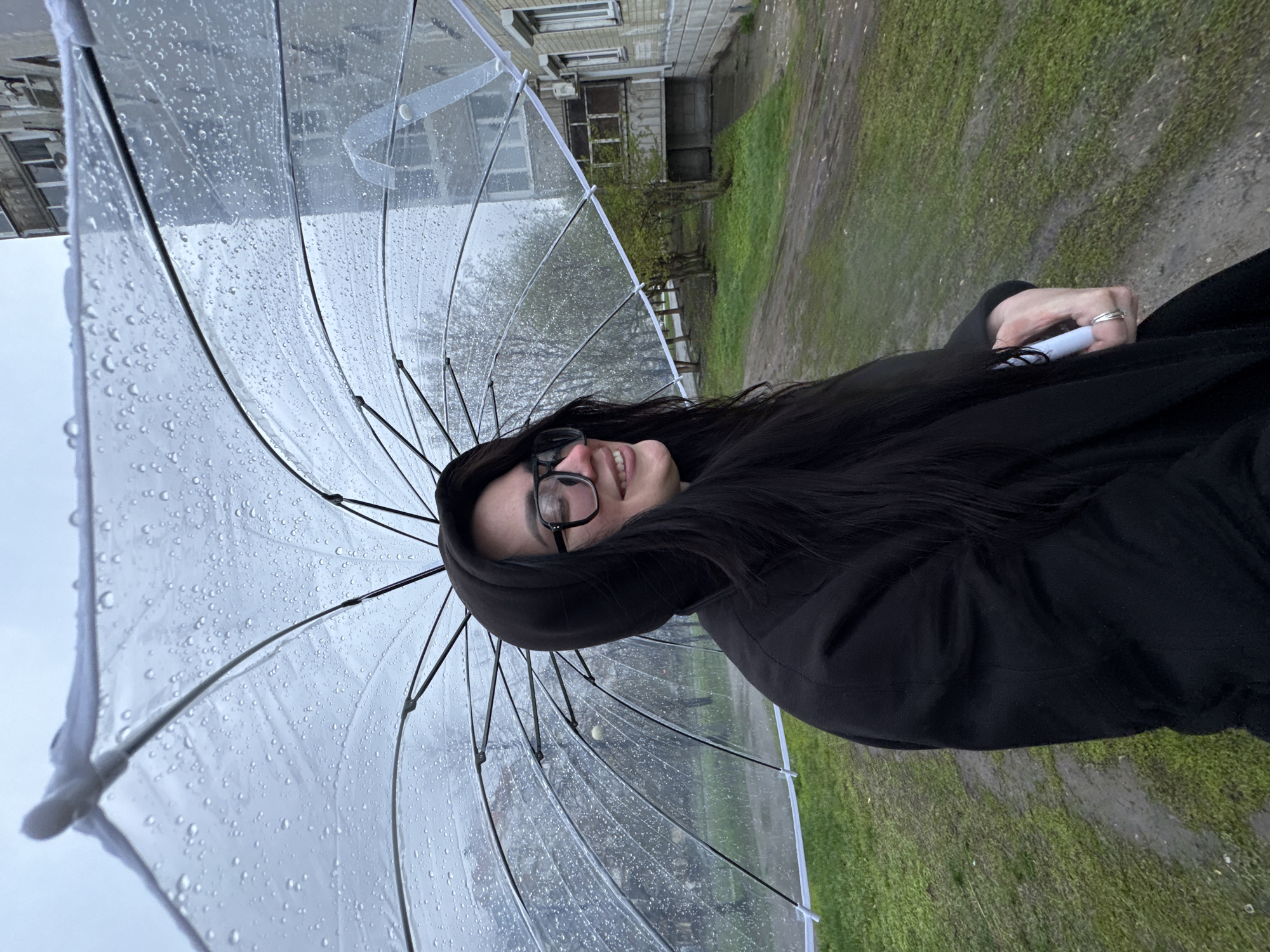 A woman stands outside under a large, clear umbrella. Raindrops are visible on the umbrella's surface. The woman is wearing glasses and a dark hooded jacket. She is smiling. In the background is a building with several windows and green grass. The scene appears to be on a sidewalk, likely in an urban area. The weather is overcast.