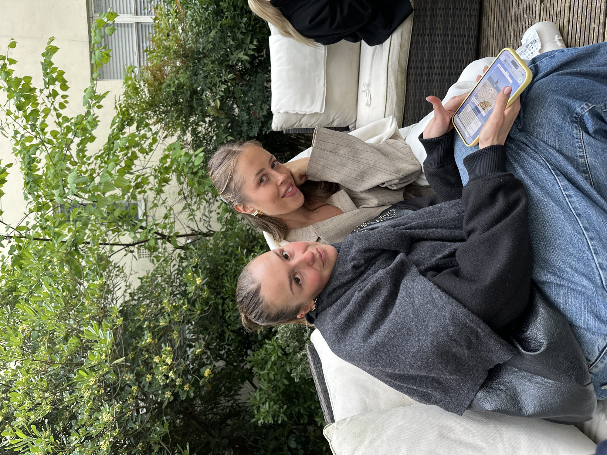 Two women are sitting outdoors on white cushioned seating in what appears to be a patio or garden area. The scene is partially framed by lush green trees and foliage. One woman is holding and looking at a smartphone with a yellow case, wearing blue jeans and layers of dark colored tops, while the other woman next to her wears a beige colored jacket and smiles. The background includes architectural elements suggesting an urban setting. The image does not show a precise location in the city of London, United Kingdom.