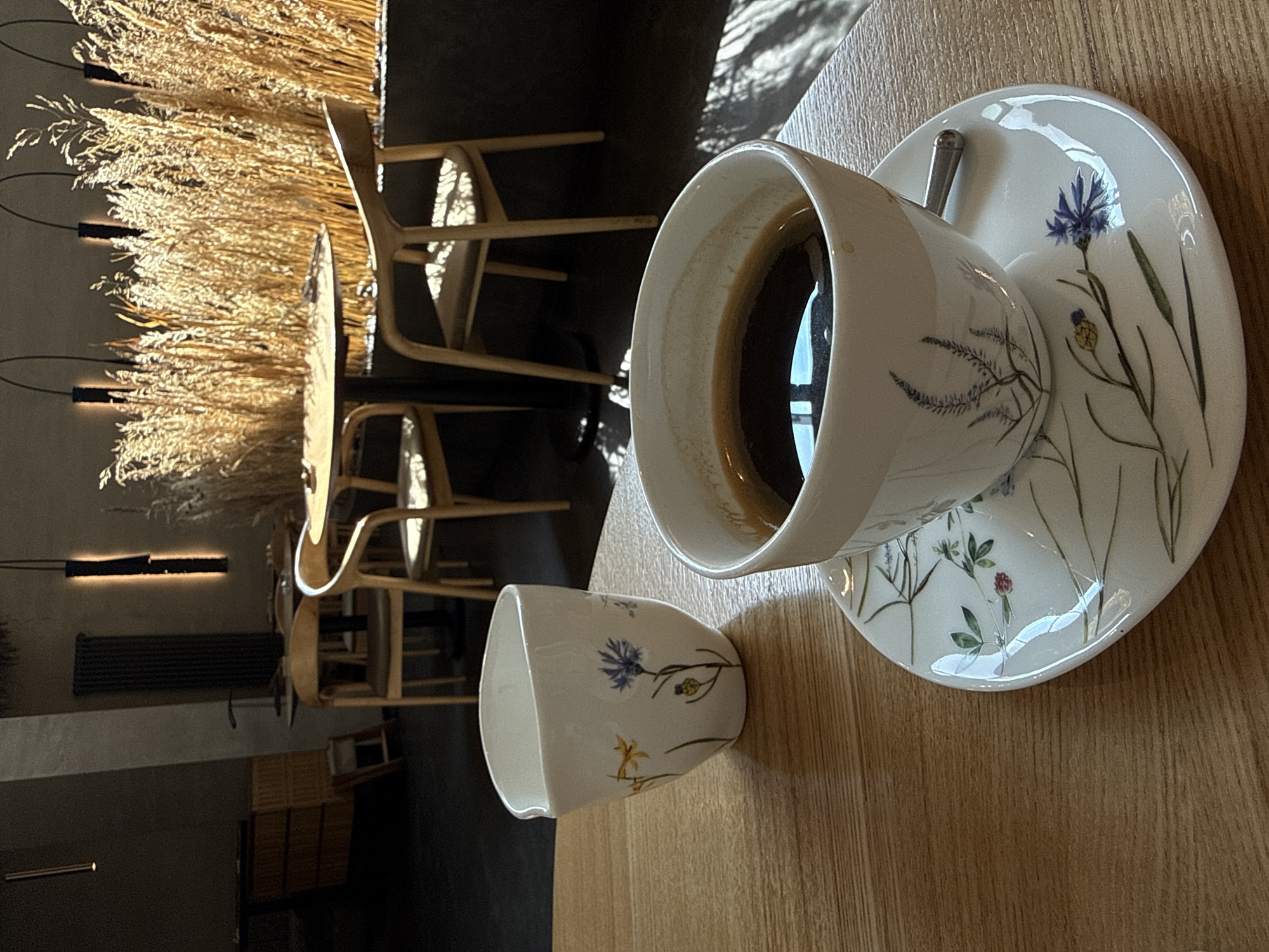 A close-up, overhead shot depicts a coffee cup and saucer, along with a smaller cup, all decorated with floral designs, sitting on a light-colored wooden table. The coffee cup is partially filled with a dark liquid, presumably coffee. A silver spoon rests on the saucer. The smaller cup is empty.
In the blurred background, there are chairs and a table arrangement. Further in the background is a decorative element comprised of golden-brown wheat stalks under warm lighting, along with architectural details such as a dark wall, ceiling lights, and a radiator.
There is no visible indication that this is the city of Krasnoyarsk, Russia.