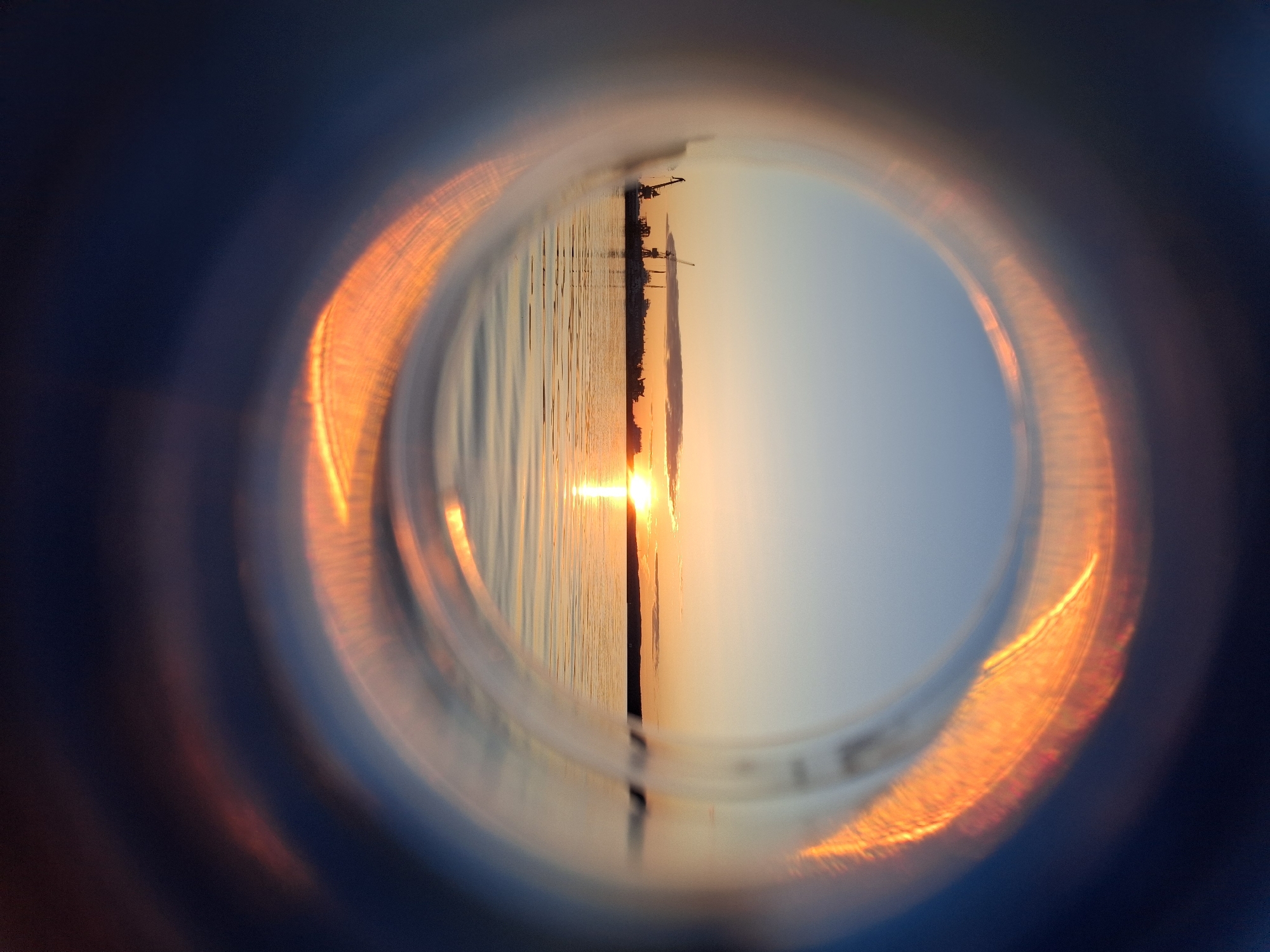 The image shows a distorted view of a sunset over water, likely a river or bay, seen through a circular lens, possibly the bottom of a plastic bottle. Silhouetted against the sunset are several cranes, indicating an industrial or port area. The water reflects the light of the setting sun. The focus of the image is inside the lens, and it's unclear what kind of camera was used to take this picture. The location is Chaikovsky, Russia, inferred from the presence of cranes and the sunset over water, aligning with its geography.