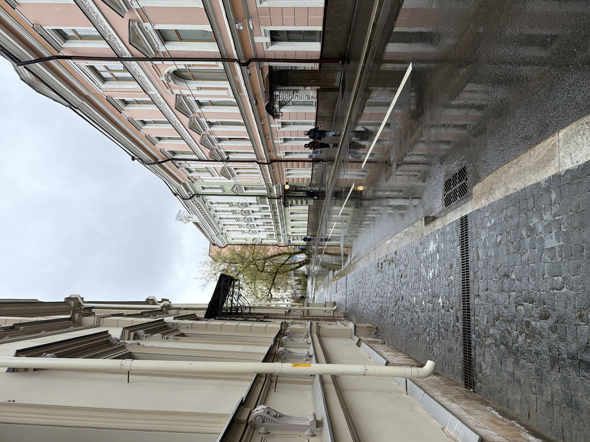 The image captures a street scene in Moscow, Russia, during or after a rain shower. The view is from a low angle, looking down the street. The street is paved with dark gray cobblestones, reflecting the wet conditions.
Buildings line both sides of the street. The building on the left has a light beige facade with ornate architectural details. A large white pipe runs horizontally along the building. The building on the right has a pink facade with white trim and numerous windows.
Several people are walking along the street, reflected in the wet pavement.
The sky is overcast and gray. A tree with sparse green leaves is visible in the middle distance. A black wrought-iron balcony is attached to the building on the left. A drain runs along the edge of the street.