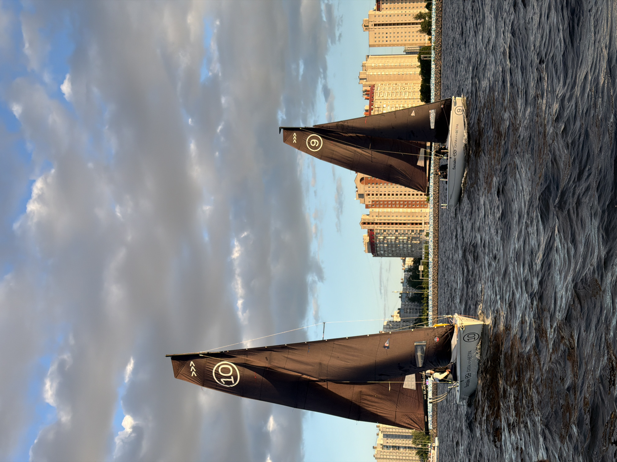 Two sailboats, one numbered 10 and the other 9, with dark brown sails featuring white symbols, are on the dark, choppy waters of the Neva River in Saint Petersburg, Russia. The sailboats are participating in a regatta, as indicated by the racing numbers. The cityscape in the background consists of modern apartment buildings, with the sun casting a warm, golden light on them, suggesting it is late afternoon or early evening. The sky is a mix of blue and dramatic clouds. On sailboat 10, a person in a white cap is visible at the helm. On sailboat 9, two individuals are seated in the cockpit. The water's surface is active, with white caps and ripples indicating a moderate wind. The overall scene depicts a sporting event against the backdrop of the urban environment of Saint Petersburg.