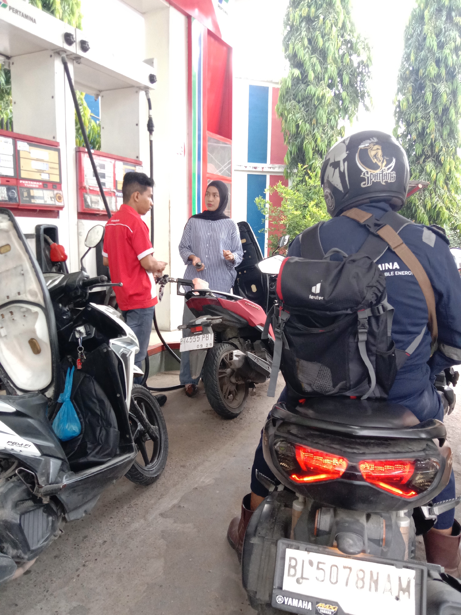 The photograph captures a scene at a Pertamina gas station. The station is identified by the Pertamina branding visible at the top of the frame. Two people, a man in a red shirt and a woman with a hijab, are standing next to a red motorcycle with license plate "BK4555 PBT 09 29". The man appears to be a gas station attendant. Another motorcycle is positioned to the left with an open seat and a blue plastic bag hanging from it. In the foreground, a person wearing a helmet with a "Spartans" logo is sitting on a black Yamaha motorcycle with license plate "BL 5078 NAM". The person has a black Deuter backpack. The environment suggests an outdoor location, with trees and buildings visible in the background. The location is Lhokseumawe, Indonesia, based on the license plate format.