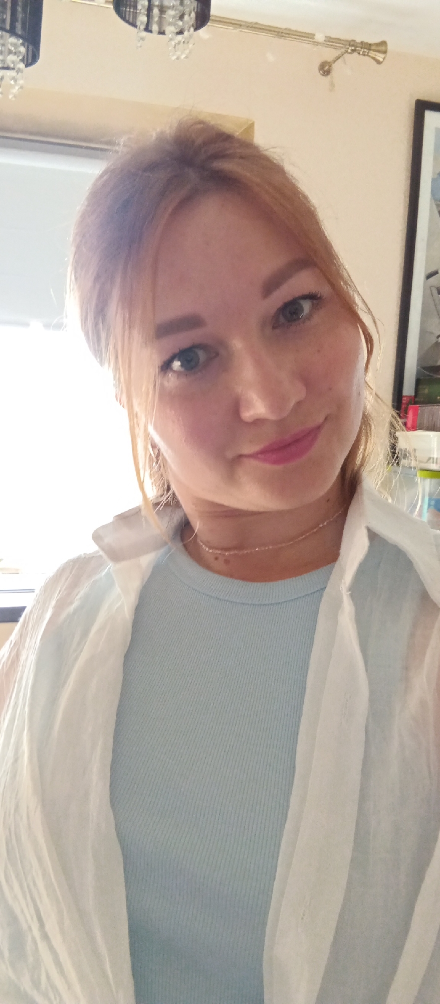 A close-up portrait captures a young woman with light reddish-blonde hair styled slightly off her face. She has fair skin with subtle freckles and a neutral yet gentle expression, her lips bearing a hint of pink. Her eyes are a light color, possibly blue or green. She is wearing a light blue ribbed top beneath a sheer white shirt, with a delicate gold chain necklace visible around her neck. The background is a soft, nondescript beige wall. A window is visible to the left. Above her, the partial view of a light fixture with crystals is visible. To her right, part of a picture frame and objects on a shelf are visible in the background. There is no indication of the city, or location within it.