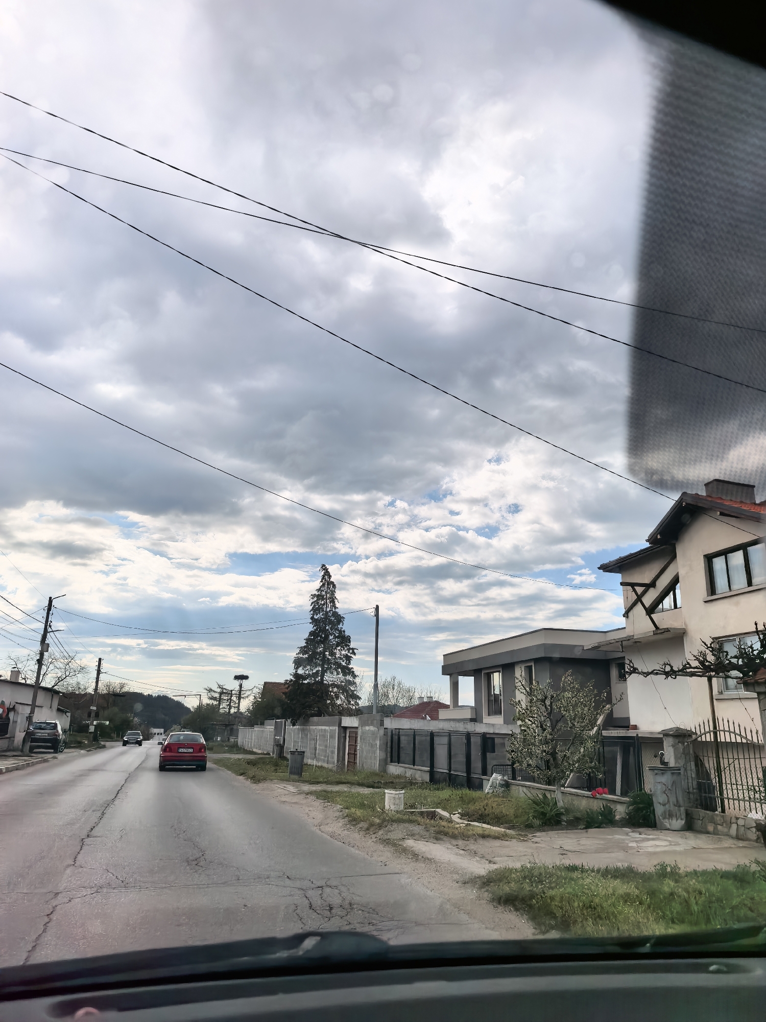 The image depicts a street scene in an urban area. The view is from inside a vehicle, looking out onto a road. A red car is traveling on the road in the same direction as the vehicle from which the image is taken. Other cars are visible in the distance. Buildings line the side of the road, including houses with varying architectural styles and fences. Power lines are strung across the sky above. The sky is overcast with a mix of grey and white clouds. Vegetation is present along the roadside.