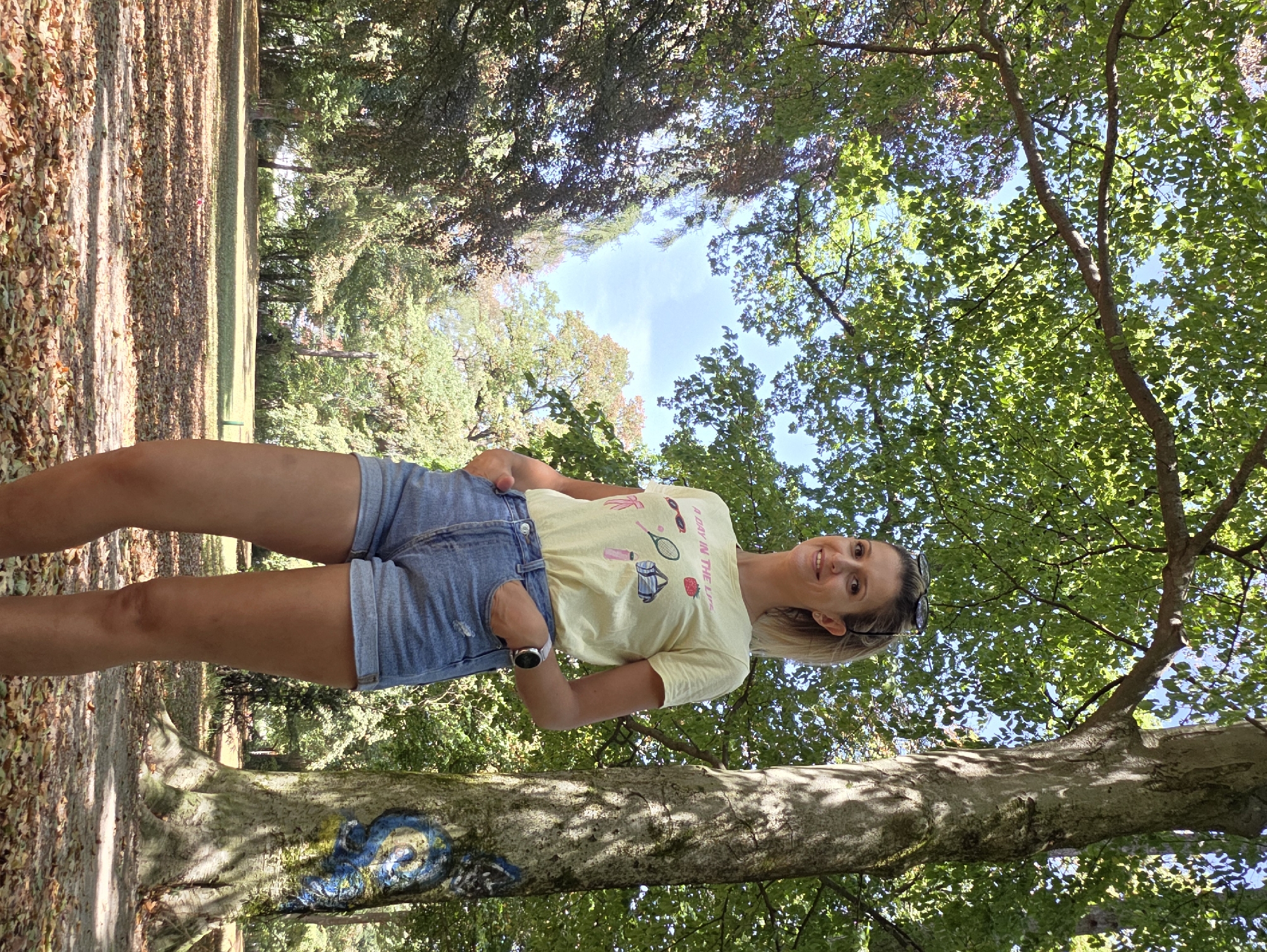 A woman with blonde hair and a bright smile poses in a park in Munich, Germany. She is wearing a yellow t-shirt with a graphic print and light blue denim shorts. Her left hand is in her pocket, and she is wearing a watch on her left wrist. Sunglasses are perched on the back of her head.
The scene is an outdoor setting with abundant greenery and trees, suggesting a daytime setting. Sunlight filters through the leaves, creating dappled shadows on the ground and on a large tree trunk in the foreground. The ground is covered with fallen leaves, indicating it might be autumn, though the lush green of the trees suggests it could also be late summer.
The woman's body is angled, and she is looking towards the camera, conveying a sense of happiness and relaxation. The background shows more trees and a clear blue sky.
The t-shirt has text on it, partially visible and in English, reading: "A DAY IN THE LIFE". There are also small graphics of a tennis racket, balls, lipstick, sunglasses, and a strawberry.