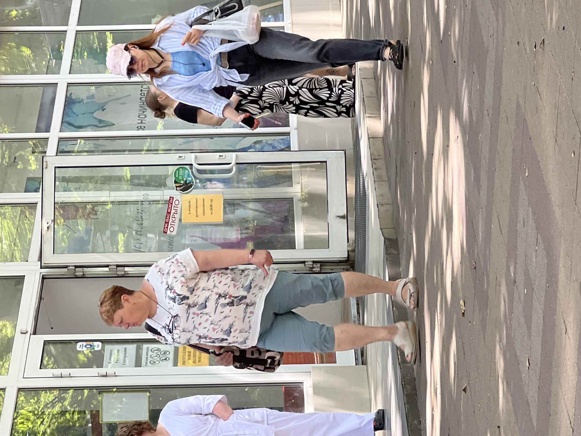 This is a medium shot, eye-level, outdoor photograph of a street scene in Krasnodar, Russia. The scene is dominated by the facade of a store with large glass doors and windows, reflecting the surrounding greenery and sky. A sidewalk runs in front of the store, with patterned paving stones.
On the left, a woman in a white uniform, possibly a nurse's or doctor's scrub suit, is partially visible, walking away from the camera. Her dark shoes are visible.
In the center, a woman with short brown hair is walking towards the right. She is wearing a white t-shirt with a colorful bird print, light blue capri pants, and light-colored sandals. She has a dark bag slung over her left shoulder and glasses hanging around her neck.
To the right of the center, a woman wearing a light blue and white striped long-sleeved shirt tied at the waist, dark jeans, a pink baseball cap, and sunglasses is walking towards the right. She has a blue tank top visible underneath her shirt and is wearing black sandals. She is holding a smartphone in her right hand and a white plastic bag containing items in her left hand. She has earbuds in her ears.
Behind these two women, partially visible, is another person in a black t-shirt and dark pants, and a person wearing a long black and white patterned skirt.
Several signs are visible on the store's glass doors. One sign, partially obscured by the central woman, appears to be in Russian and indicates "OPEN." Another sign, to the right, also in Russian, features the text "ОТ СЕБЯ PUSH" (Push from yourself). Yellow signs with Russian text are also visible, one stating "В ПРОДАЖЕ ЕЖЕДНЕВНИКИ НА 2026 ГОД" (For sale diaries for the year 2026).
The lighting suggests it is daytime, with dappled sunlight casting shadows from trees onto the sidewalk. The overall environment appears to be a commercial or public area in a city.