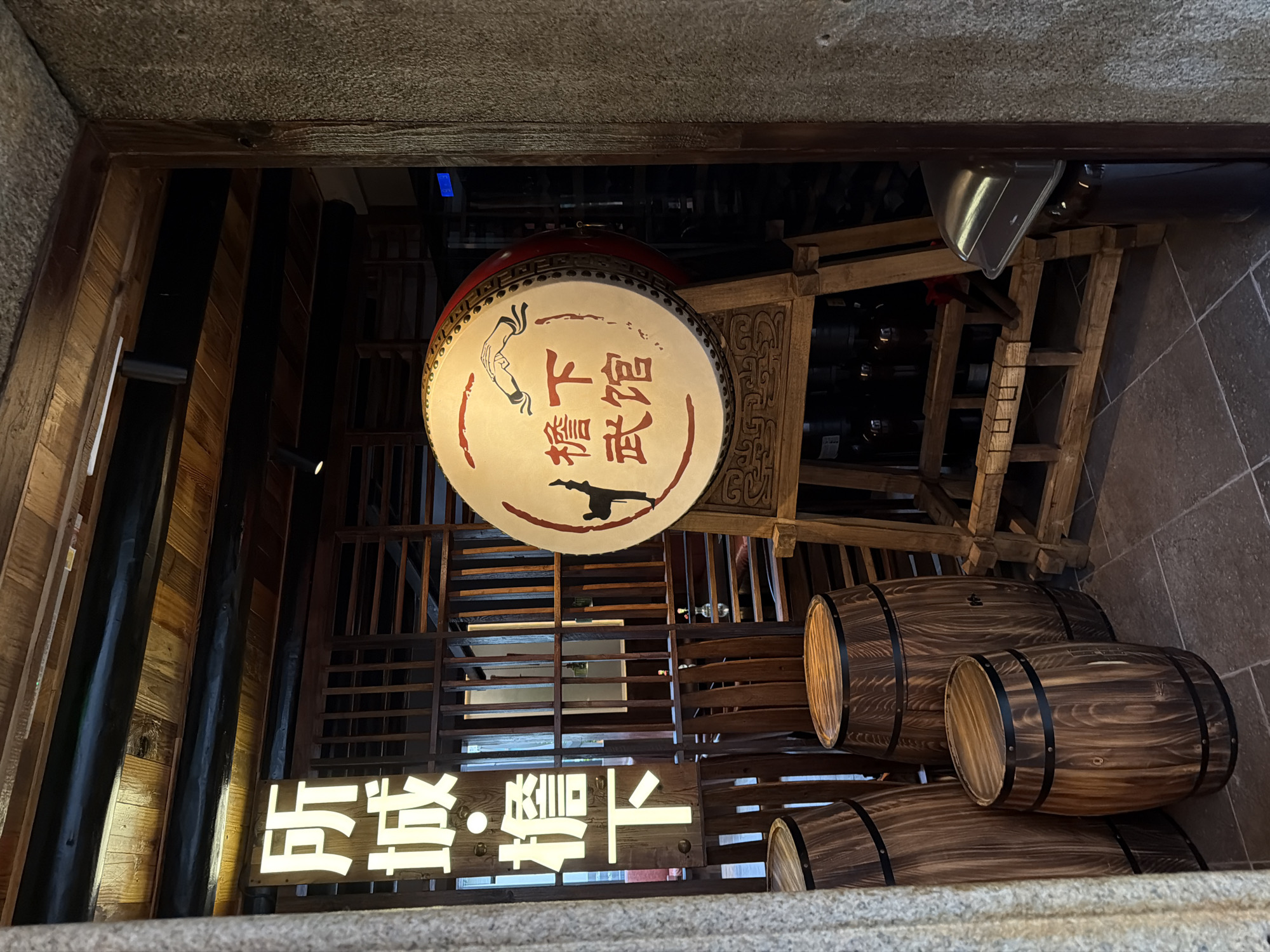 An indoor scene captures a traditional Chinese aesthetic, likely within the Longgang District of China. The foreground features three wooden barrels of varying sizes, weathered with dark bands around their circumferences. Behind them, a large, round drum, illuminated by ambient light, is prominently displayed on a wooden stand. The drum's surface bears red Chinese characters and a depiction of two hands and a bird. To the left of the drum, a vertical sign with bright white Chinese characters is mounted on a wooden lattice structure. The background reveals more wooden lattice work, a shelf system, and hints of dark bottles. The overall impression is one of a cultural establishment, possibly a restaurant or a shop, with a focus on traditional Chinese elements.