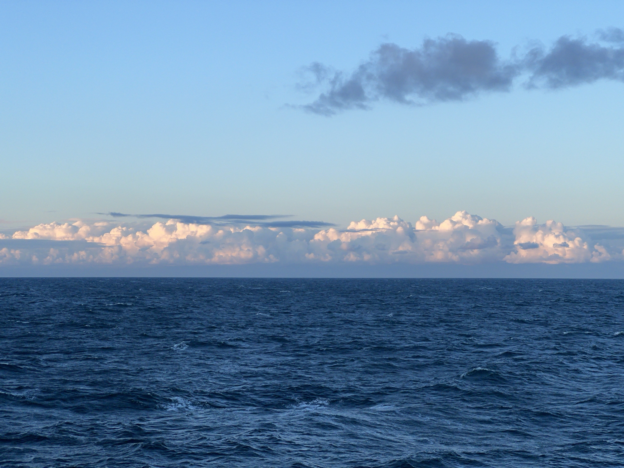 The image depicts a serene seascape under a pale blue sky. The horizon line is clearly defined, separating the vast expanse of the deep blue ocean from the sky above. White and pinkish clouds are scattered across the sky, with a few darker clouds visible in the upper portion. The ocean surface is rippled, suggesting a gentle breeze. The overall impression is one of natural tranquility, with no human presence or man-made structures visible. The location is identified as Unknown, Poland.