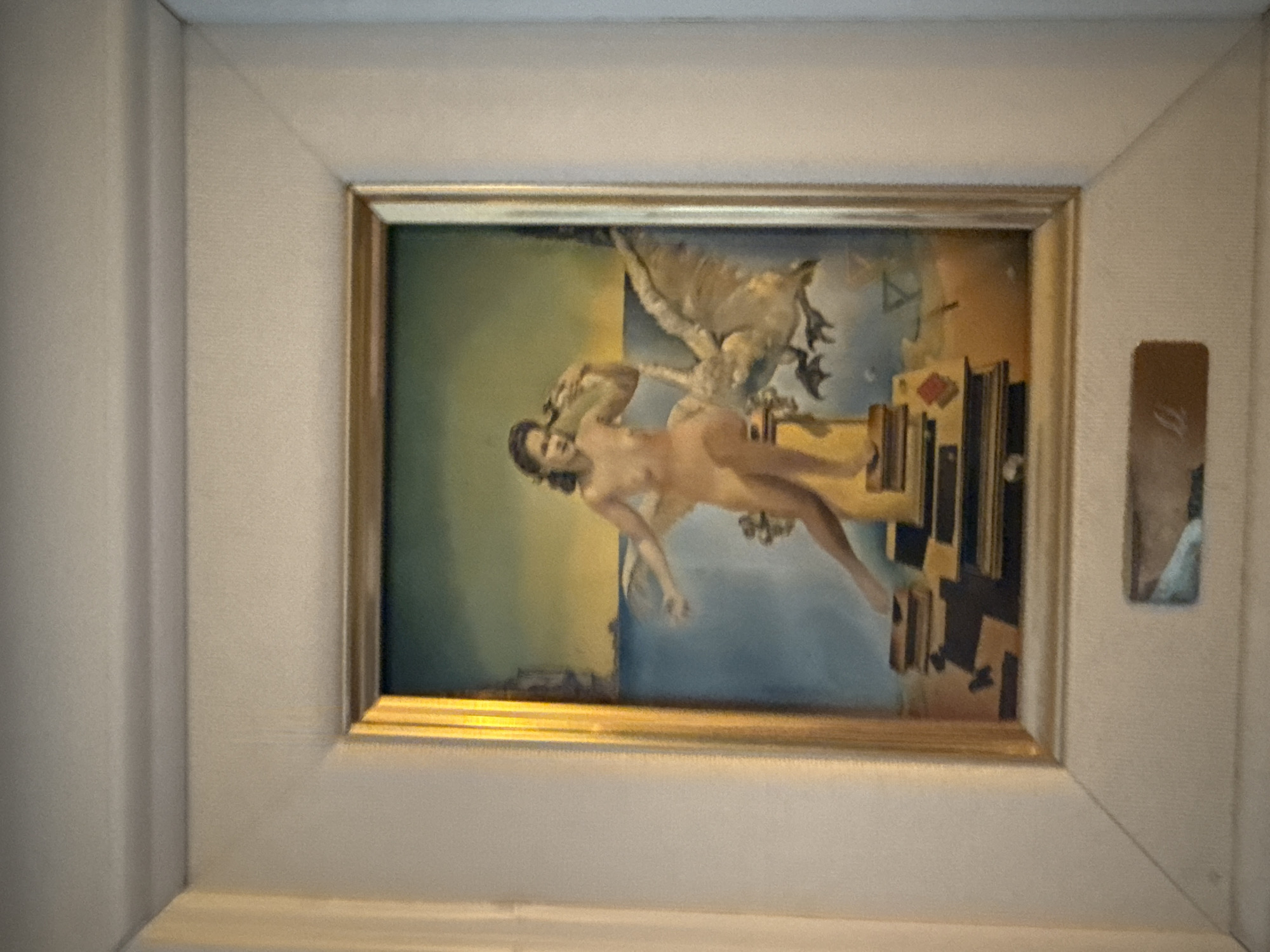 The image shows a framed surrealist painting featuring a nude woman and a large bird against a dreamlike landscape. The woman, with dark hair and a fair complexion, is positioned on a stack of books or platforms. She is holding a bird or a similar object in her right hand and her left arm is extended forward. To her right, a large, feathered bird with its wings spread appears to be flying or swooping down. The background depicts a body of water, a distant coastline with structures, and a gradient sky transitioning from blue to yellow. The painting is enclosed in a golden frame, which is then placed within a larger, white matting and frame. A small, rectangular plaque is affixed to the bottom of the white matting, featuring some indistinct lettering. The overall presentation suggests the artwork is displayed on a wall, likely in a domestic or gallery setting. The image does not contain any information that would allow for identification of its location in the city of Yukki, Russia.