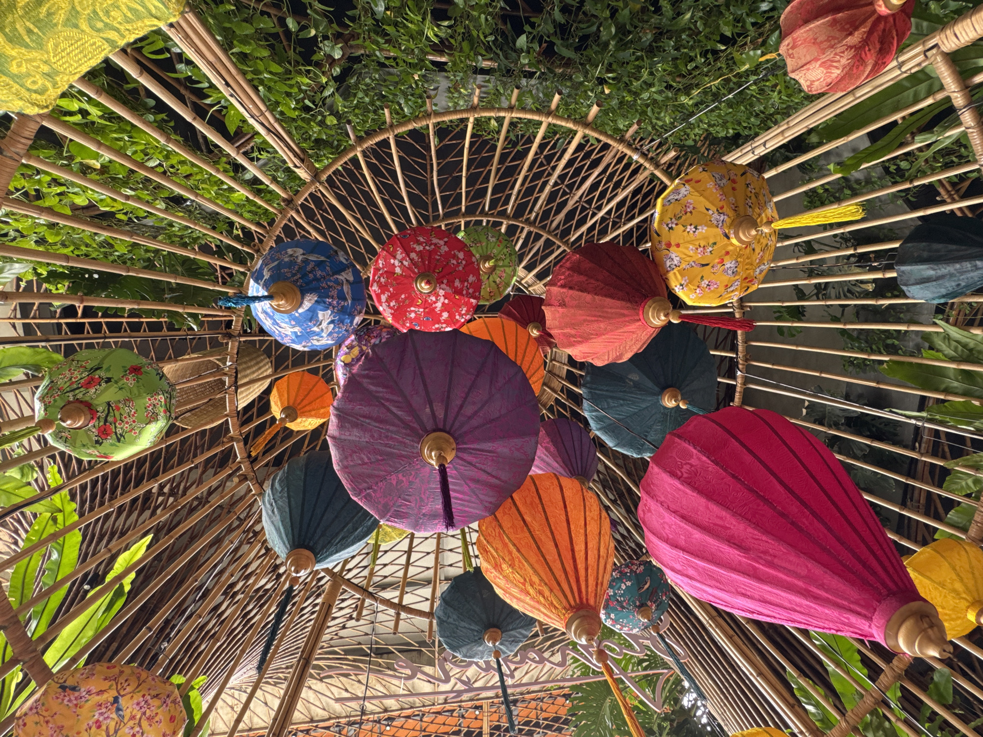 This image captures a ceiling adorned with numerous decorative lanterns in a city setting in Nha Trang, Vietnam. The lanterns are of various colors, including yellow, green, blue, purple, orange, red, and pink. They are suspended from a bamboo-like structure that forms a radial pattern. Some lanterns are plain, while others feature floral or bird patterns. The background consists of lush green foliage, suggesting an outdoor or semi-outdoor space with abundant plant life. The perspective is from below, looking up at the lanterns and the ceiling structure. There are no people visible in the image.