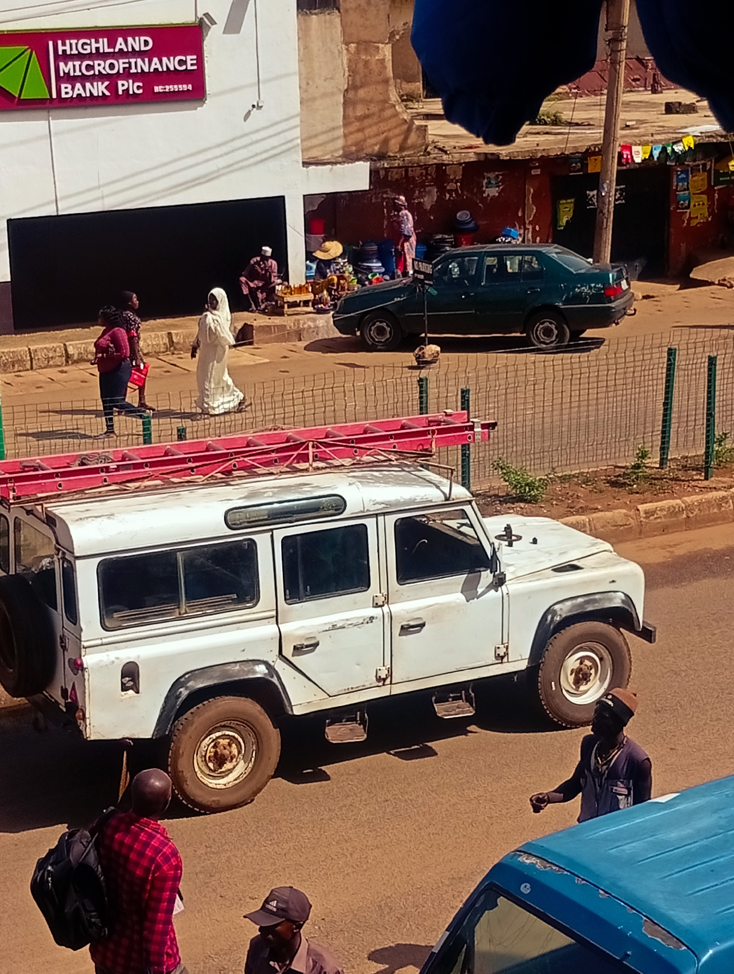 In Bukuru, Nigeria, on a sunny day, a white Land Rover Defender with a red ladder strapped to its roof is parked on a street. In the foreground, two men stand near the vehicle. One man, wearing a red and black plaid shirt and a black backpack, faces away from the camera. The other man, wearing a dark vest and a cap, faces the Land Rover. Behind the Land Rover, a green sedan is parked on the street, and a chain-link fence separates the street from a sidewalk. On the sidewalk, three individuals are visible: a person in a white garment, a person in a pink top and blue jeans, and another person with their back to the camera. A building with a sign that reads "HIGHLAND MICROFINANCE BANK Pic" is in the background. The ground is a mixture of asphalt and dirt.
